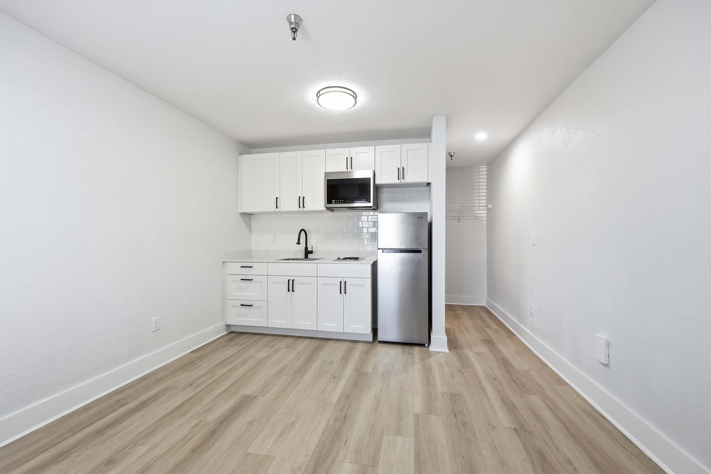 A small, modern kitchenette with white cabinets, a stainless steel refrigerator, and a microwave. The space features light wood flooring and white walls, creating a bright and clean atmosphere. There is a sink and minimal counter space, with an area for storage in the background.