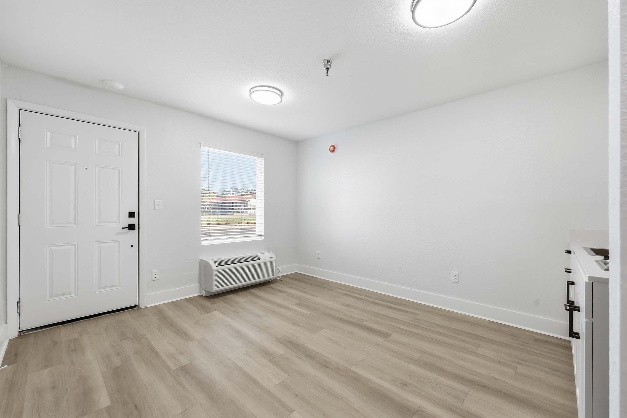 Empty room with light-colored walls and a large window allowing natural light. The floor is light wood laminate. There is an air conditioning unit on the floor and a white door leading outside. Ceiling lights provide illumination. The space is clean and minimalistic.