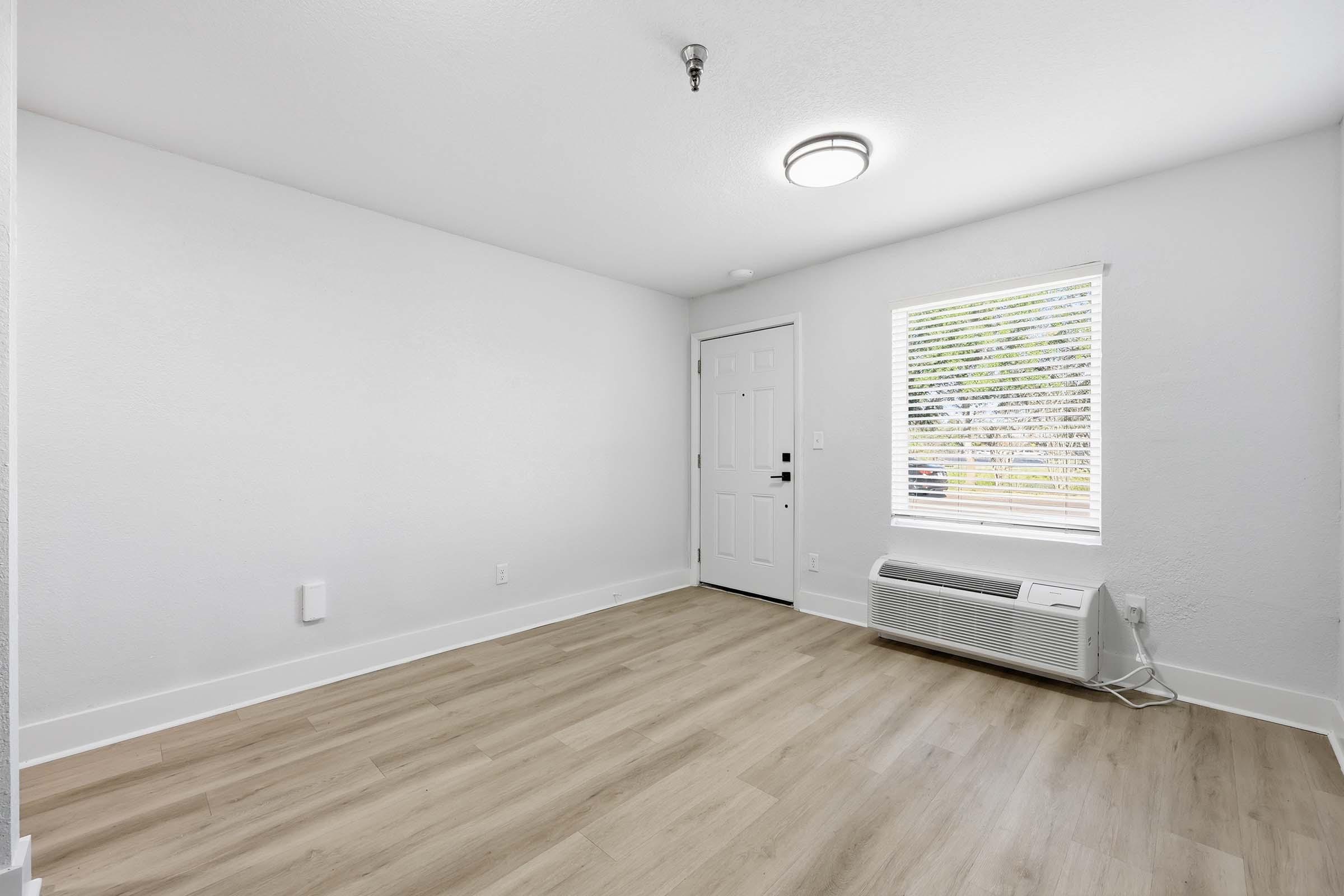A bright, empty room featuring light wood flooring and white walls. A window with blinds allows natural light, and an air conditioning unit is visible on the floor. The main entrance door is in the corner, leading to the outside. The ceiling has a modern light fixture.