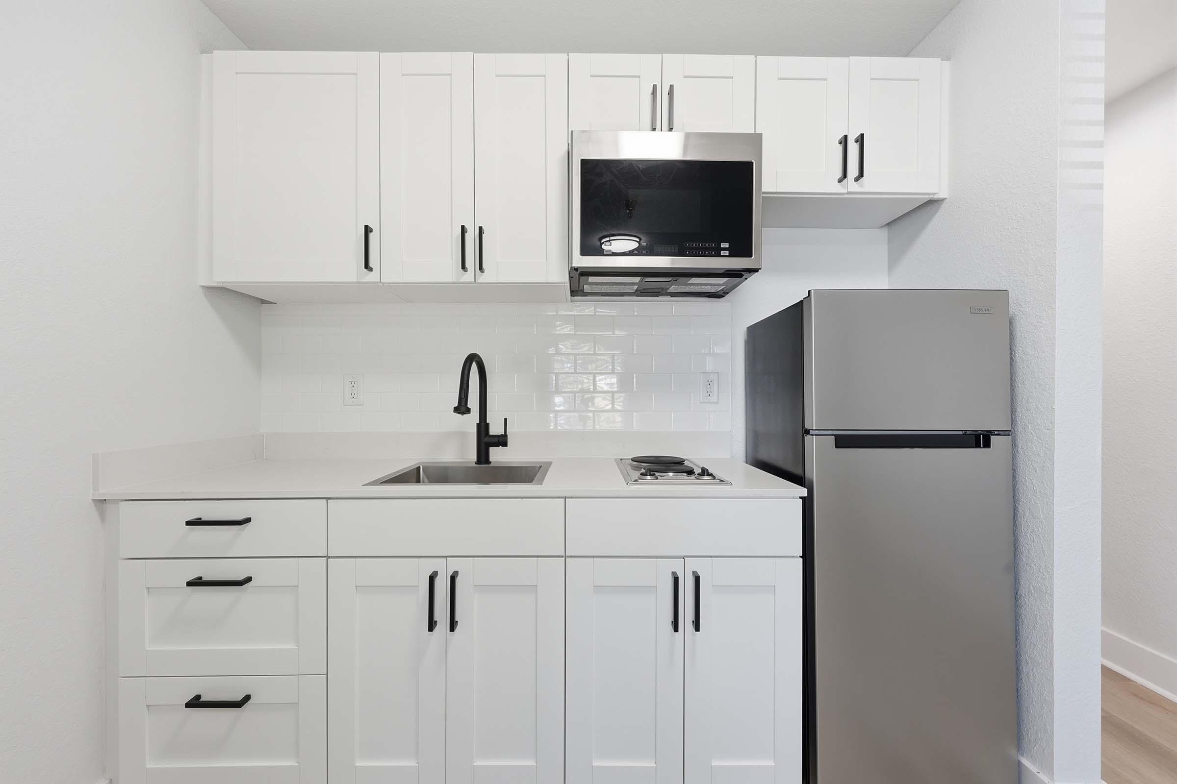 Modern kitchen with white cabinetry, a stainless steel microwave, a sleek black faucet, and a compact refrigerator. The countertop is light-colored, and there is a gas stove with a minimalist design. The overall look is clean and contemporary, featuring a bright and airy atmosphere.