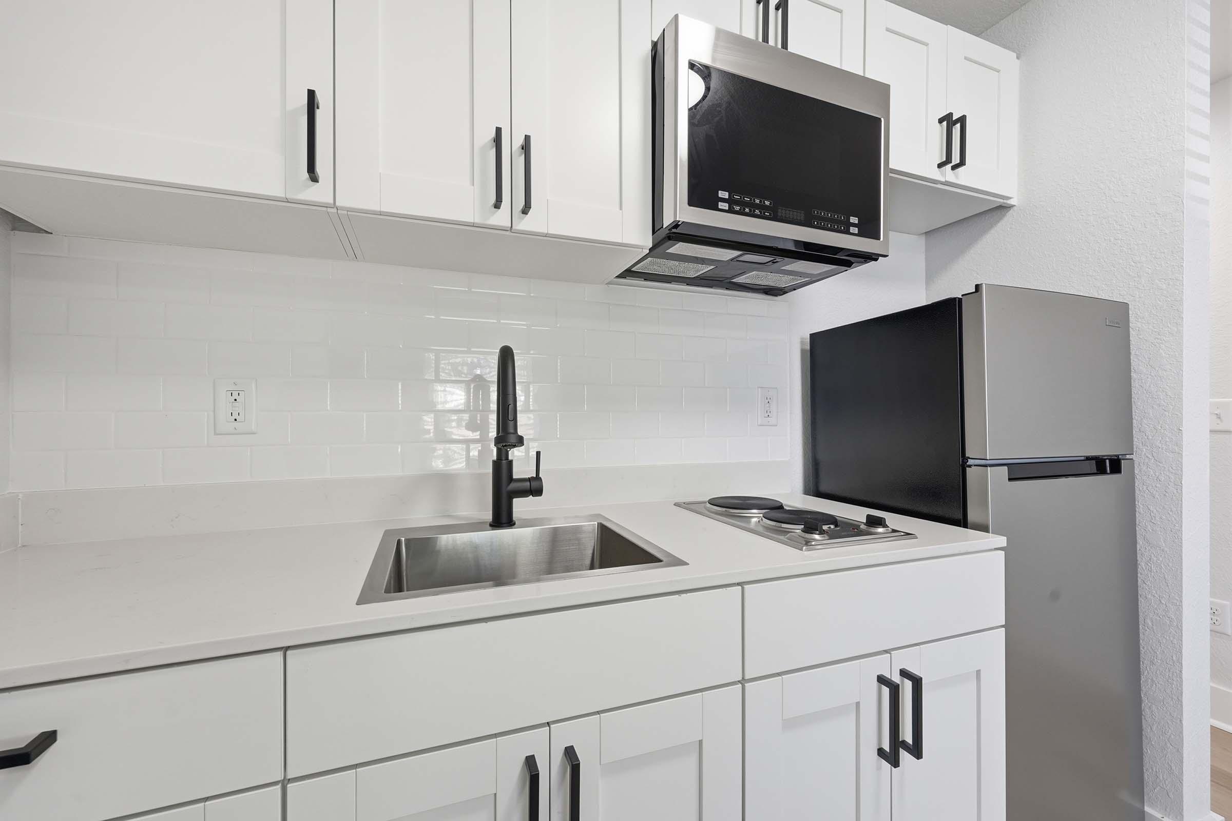 Modern kitchen featuring white cabinetry, a stainless steel microwave, a black refrigerator, a sink with a sleek black faucet, and a stovetop. The backsplash has white subway tiles, and the overall design is clean and contemporary.