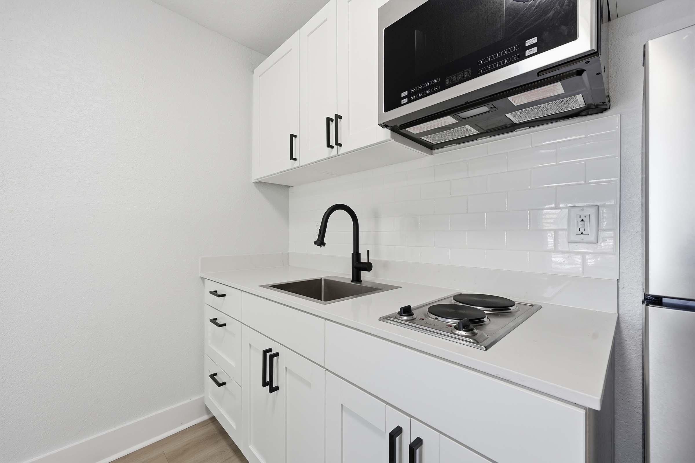 A modern kitchenette featuring white cabinets with black handles, a stainless steel refrigerator, a sink, and a microwave mounted above a compact stovetop. The backsplash is white tiled, creating a clean and bright aesthetic. The countertops are smooth and light-colored, enhancing the contemporary design.