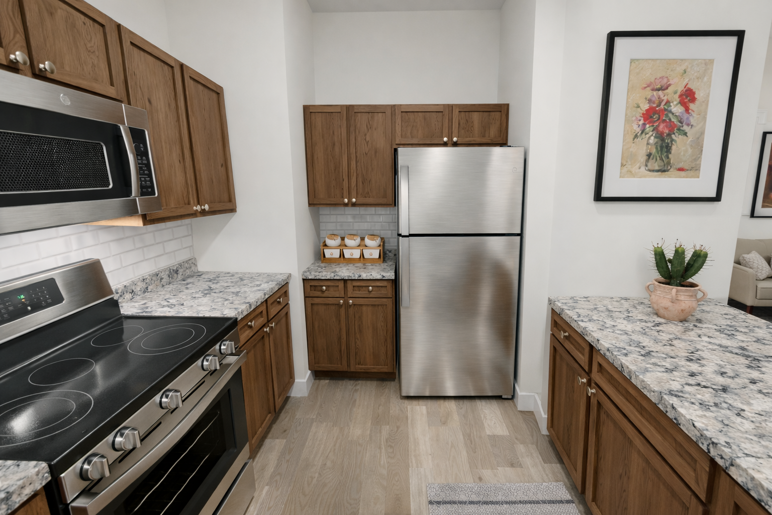 Modern kitchen featuring wooden cabinets, stainless steel refrigerator, and oven. The countertops are gray with a subtle pattern. A potted plant and a framed floral artwork are on the countertop. The space is well-lit, with an open layout showcasing the kitchen design.