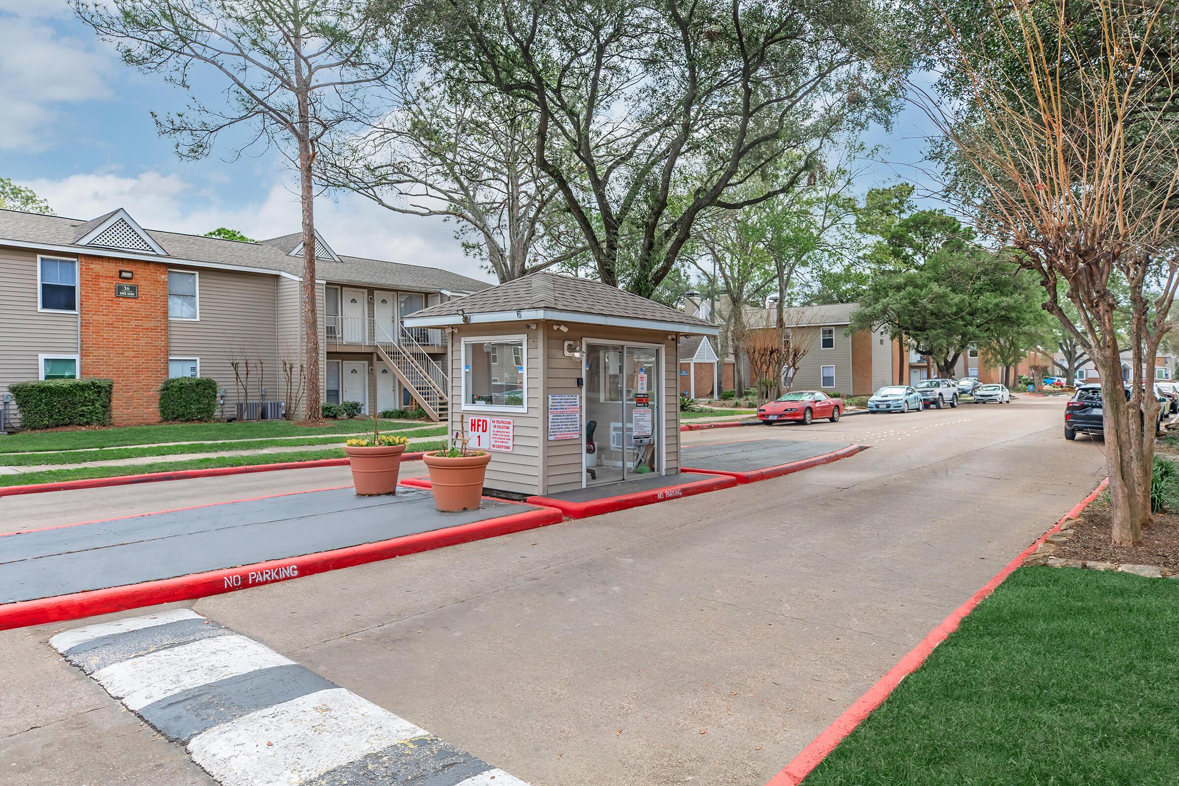 A view of an apartment complex entrance with a security kiosk on the left side, surrounded by landscaped areas. The road is marked with a red curb, and there are potted plants near the kiosk. Several vehicles are parked along the street, and multiple buildings are visible in the background.