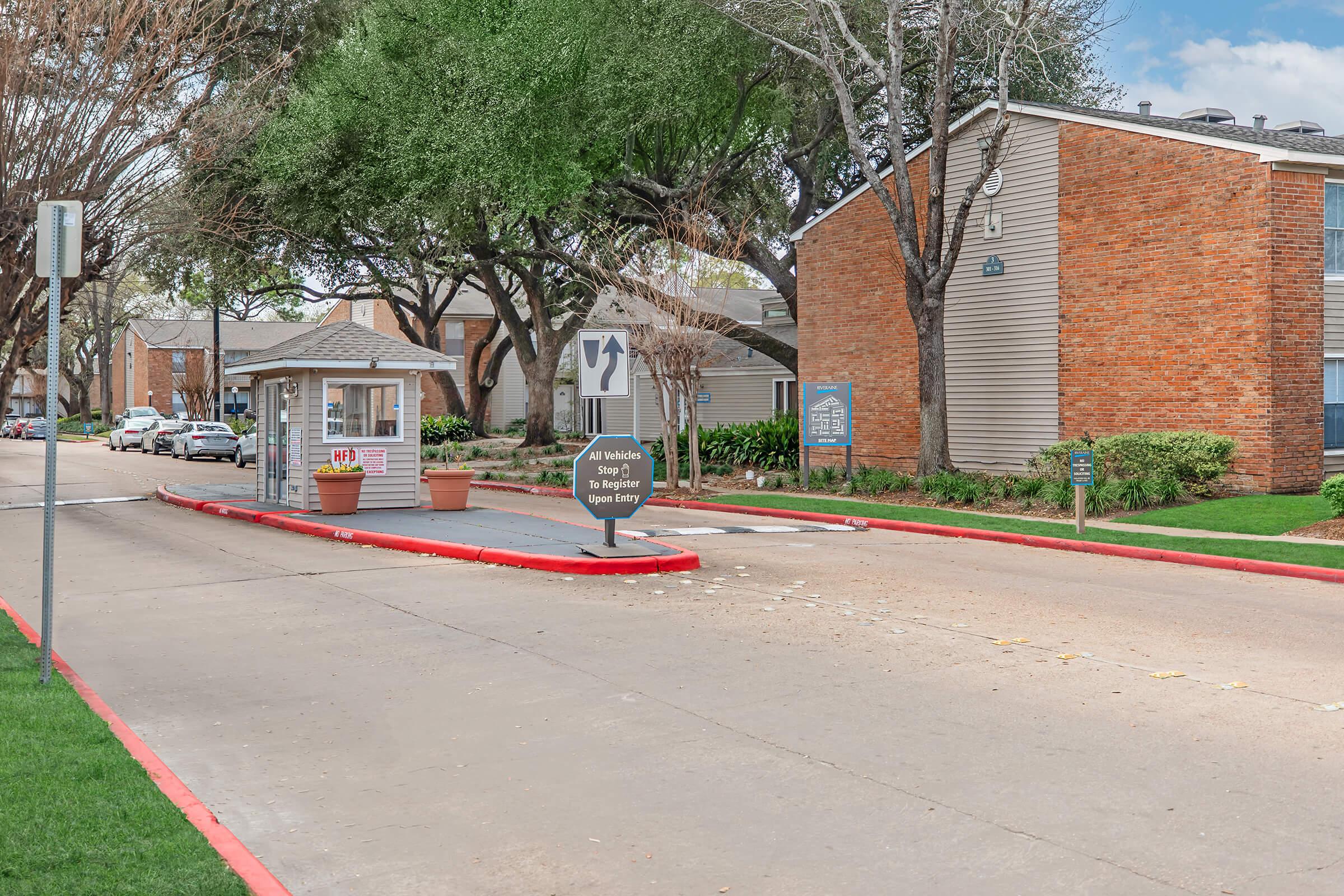 A view of an apartment complex entrance with a residential booth on the left, a directional sign indicating a left turn, and well-maintained landscaping. In the background, several apartment buildings are visible, along with trees lining the pathway. The street is paved and includes a parking area.