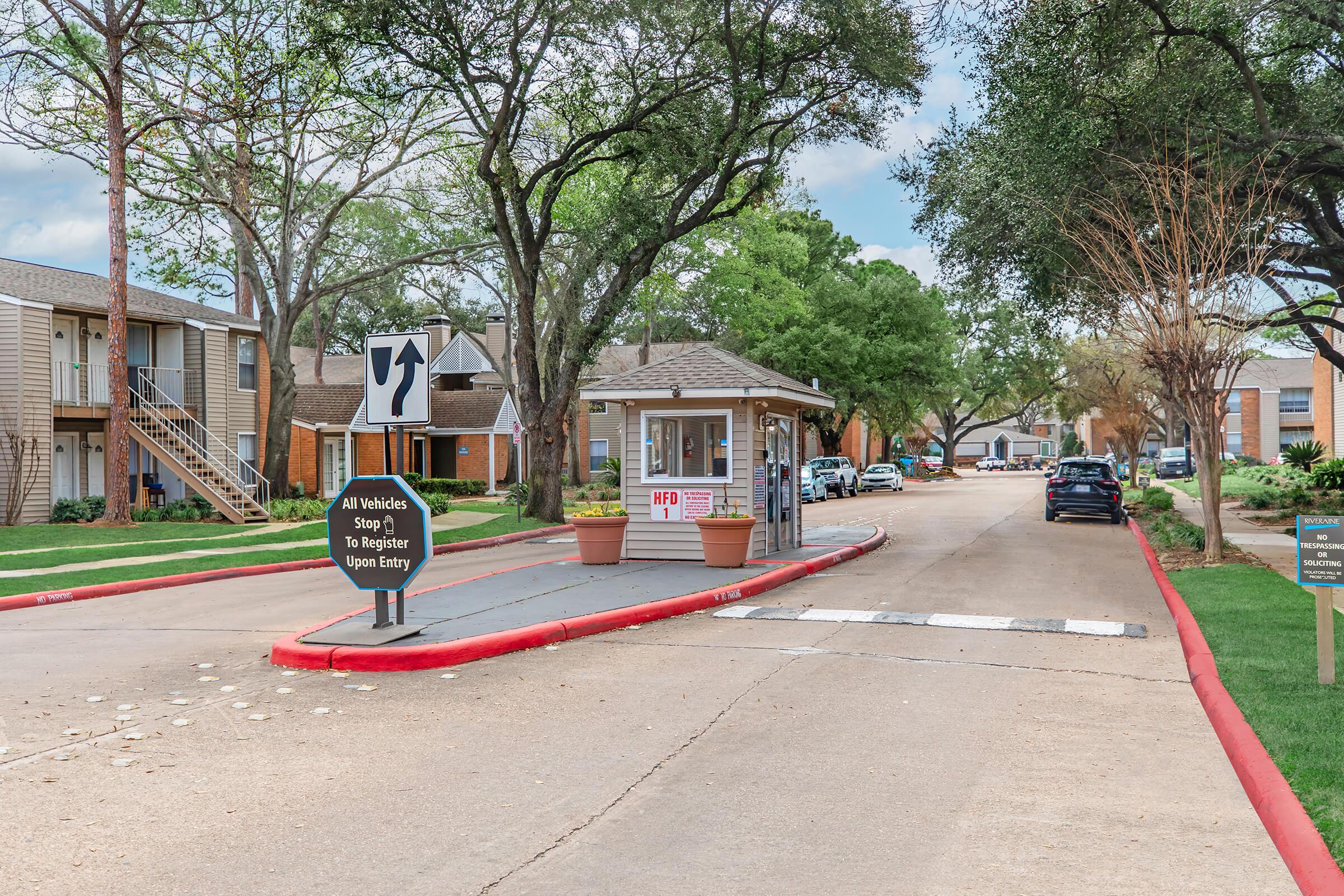 A view of an apartment complex entrance featuring a guard booth with a small sign. The road is lined with trees and greenery, and there are parked cars along the sidewalk. A directional sign indicates a left turn, and there is a notice for vehicles to stop and register upon entry.
