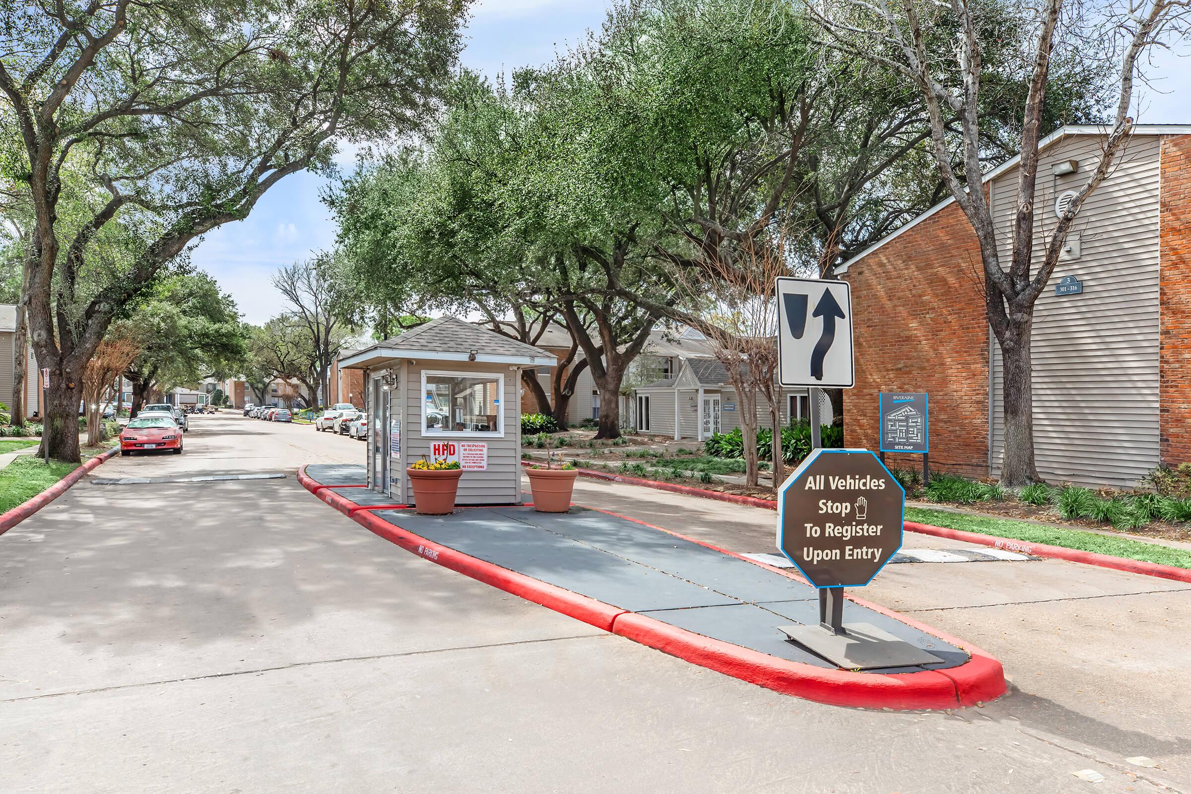 A parking entry booth located along a residential street lined with trees. A sign directs vehicles to stop and register upon entry. The booth is at the end of a paved path, with colorful flower pots nearby and apartment buildings visible in the background.