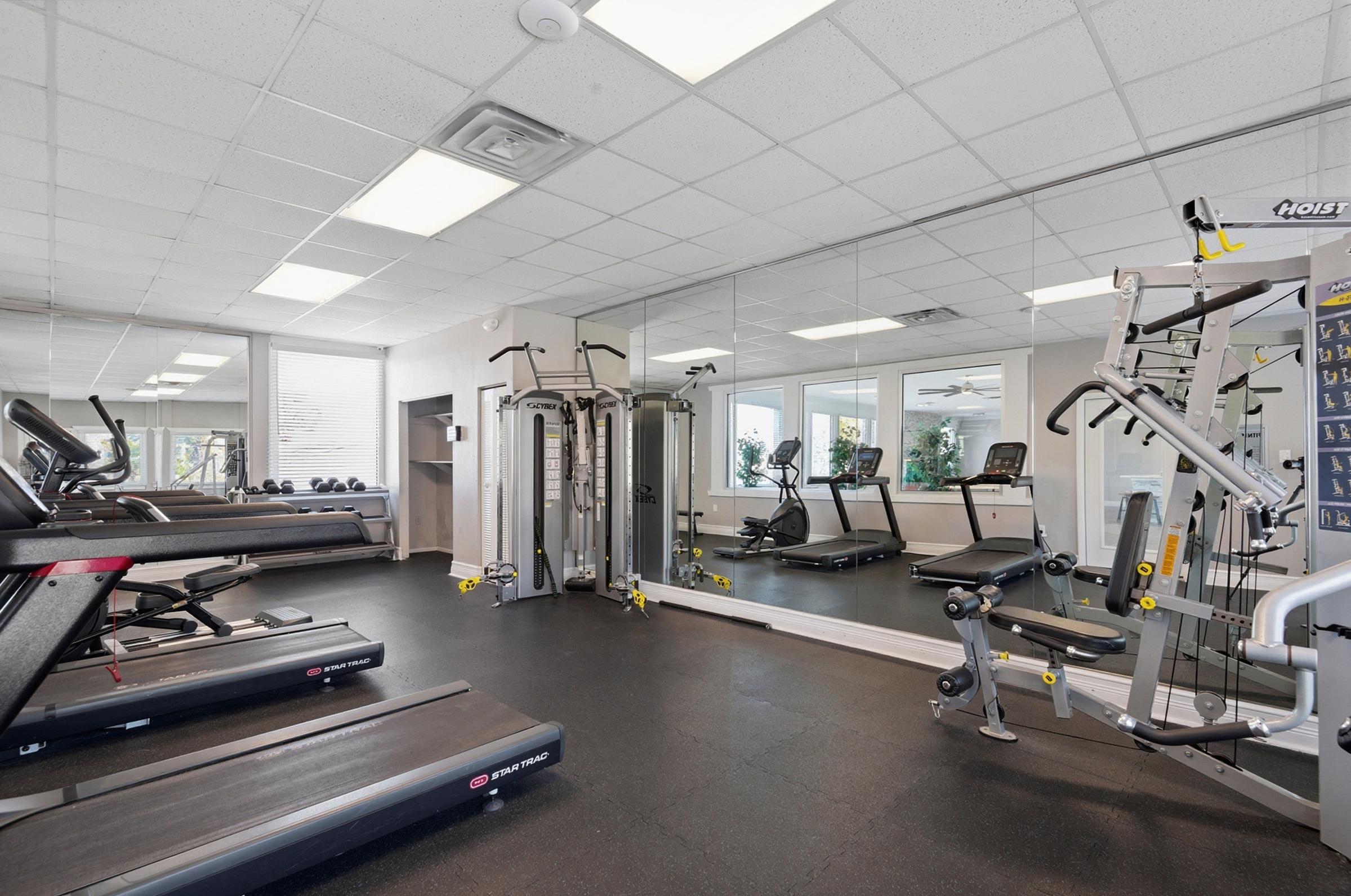 A bright, modern gym featuring various exercise equipment, including treadmills and weight machines. Large mirrors line one wall, reflecting the space. The room is well-lit with natural light coming from windows and has a clean, organized appearance, creating an inviting atmosphere for workouts.