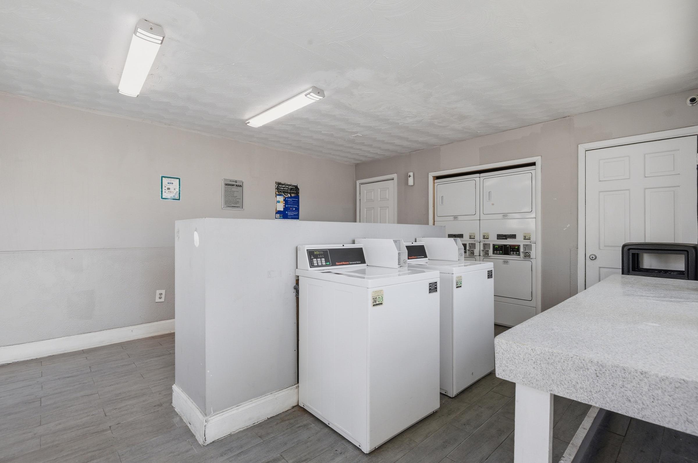 A clean and bright laundry room featuring two white washing machines and a white countertop. There are doors and appliances in the background, along with bright overhead lighting. The walls are a light gray color, creating a simple and functional space for laundry.