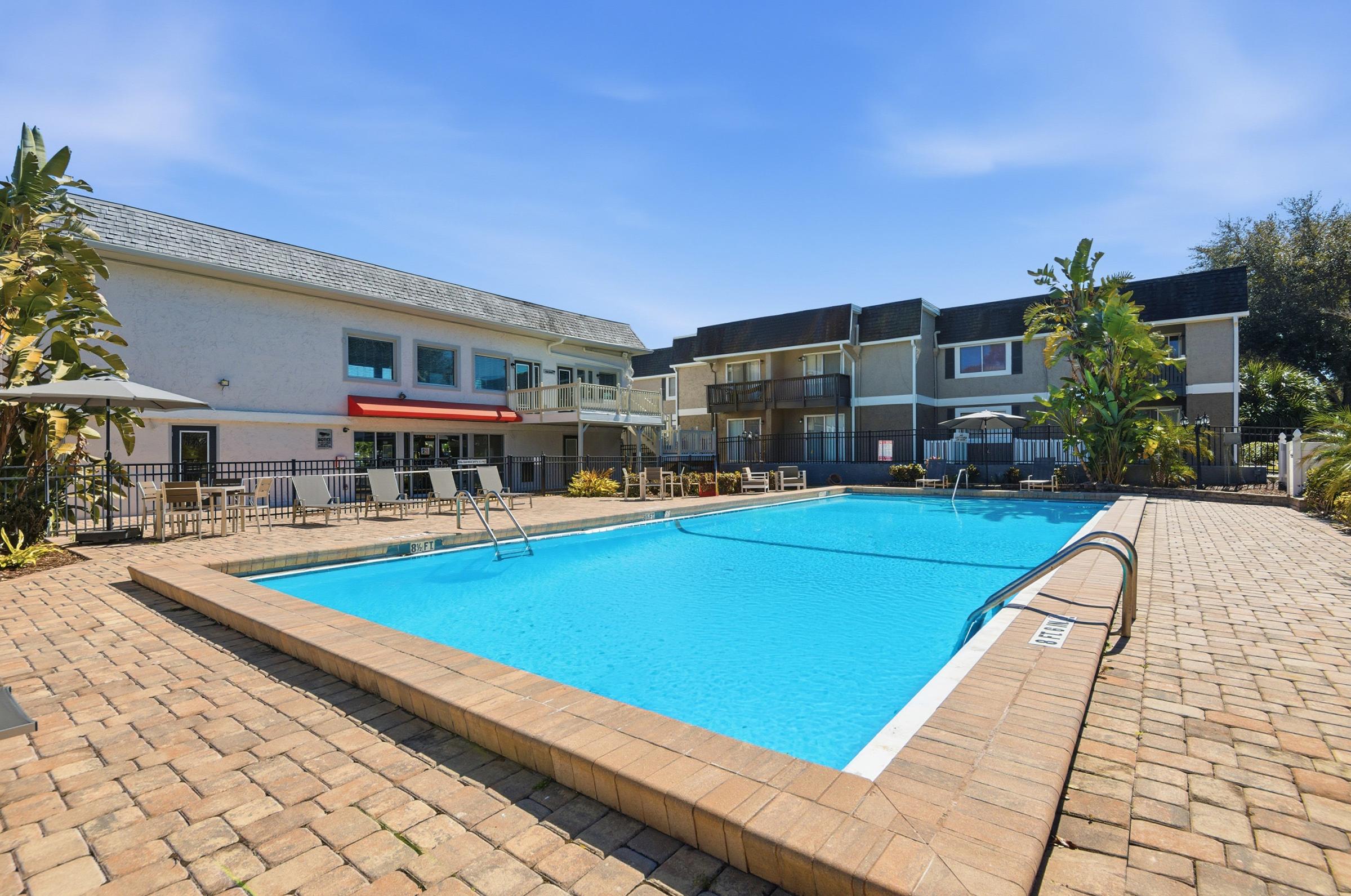A clear, inviting swimming pool surrounded by a brick patio. Lush green plants and palm trees line the area. Nearby, there are lounge chairs and umbrellas for shade. Two buildings with balconies are visible in the background, providing a serene atmosphere for relaxation.