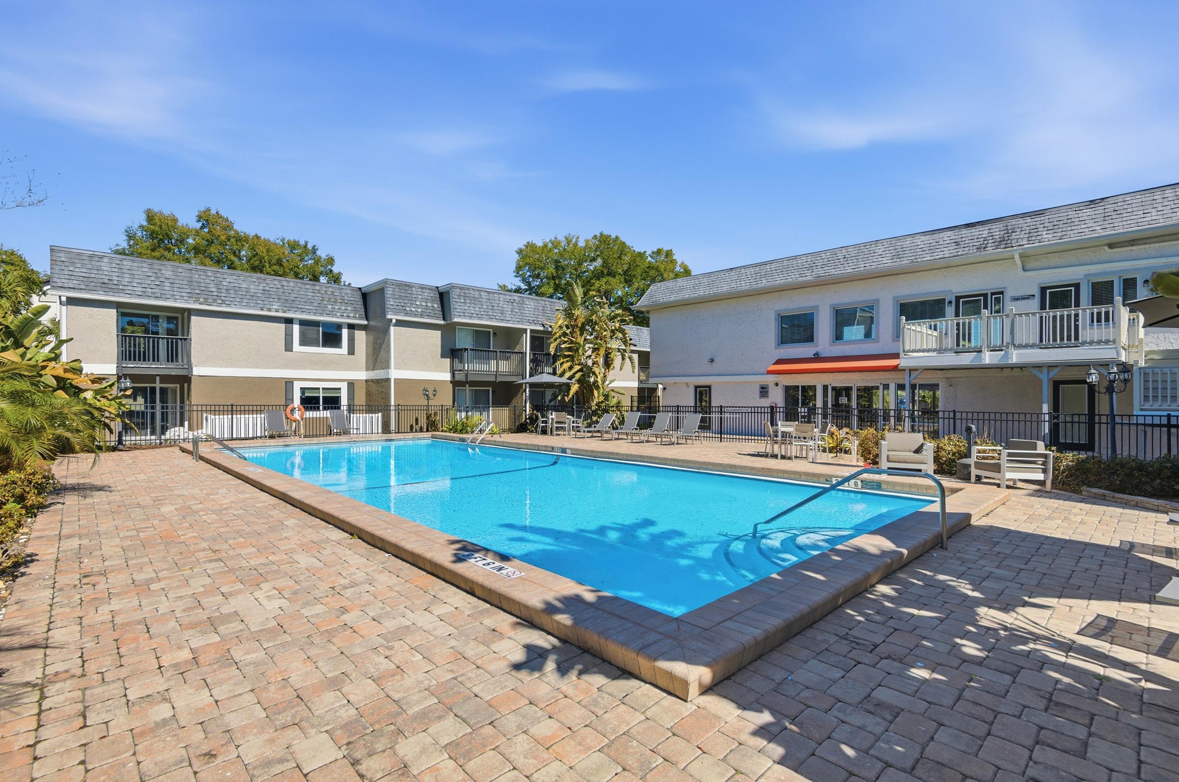 A clear swimming pool surrounded by a paved area with lounge chairs, featuring two buildings in the background. The scene is sunny with a blue sky, and lush greenery nearby, creating a relaxing atmosphere. Ideal for leisure and recreation, especially in a residential setting.
