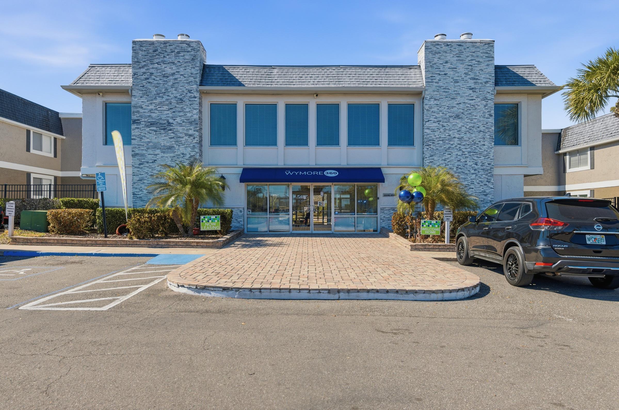 A modern building with a light blue stone facade, featuring large windows and a blue awning displaying the name “Wynmore.” There are palm trees and decorative greenery in front, along with a parking lot. A black SUV is parked nearby. Clear blue sky above.