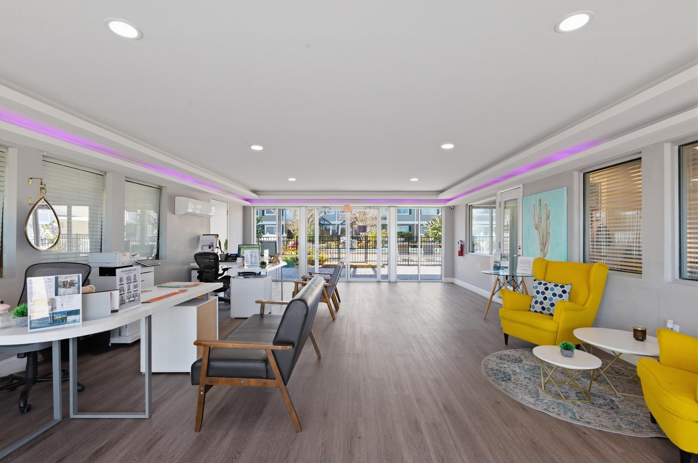 A modern office reception area featuring a bright interior with large windows. There are two seating areas: one with a yellow armchair and a round coffee table, and another with gray chairs. A desk with office supplies and a computer is visible. Soft purple lighting accents the ceiling.