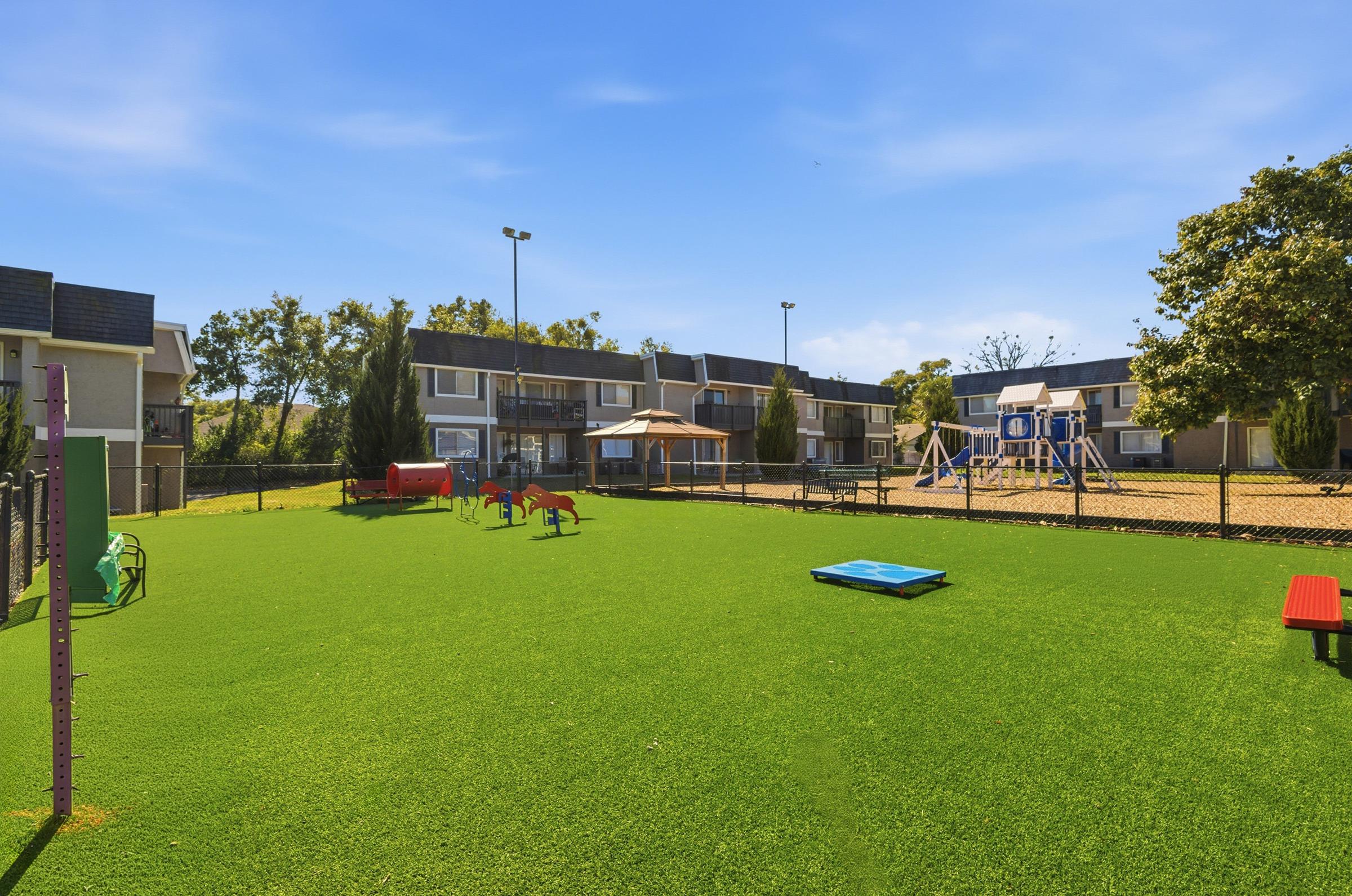 A sunny playground area featuring artificial turf, with colorful play structures such as slides and climbing equipment. Surrounding the playground are apartment buildings, trees, and a bright blue sky, creating a cheerful outdoor environment for children.