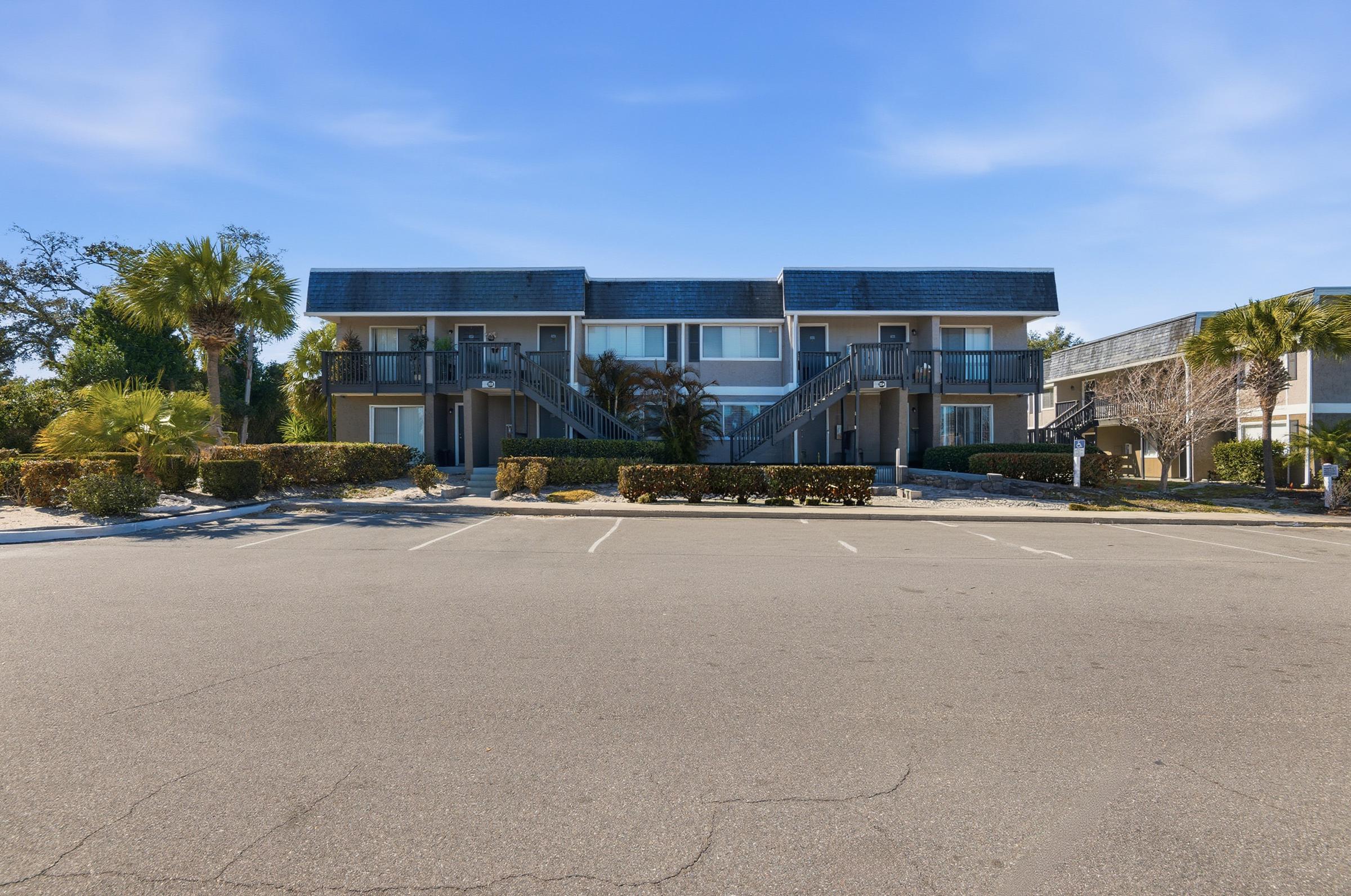 Two-story apartment complex with a modern design, featuring a slate roof and balconies. Surrounded by manicured landscaping, palm trees, and parking spaces. A clear blue sky provides a bright backdrop, creating a welcoming atmosphere.