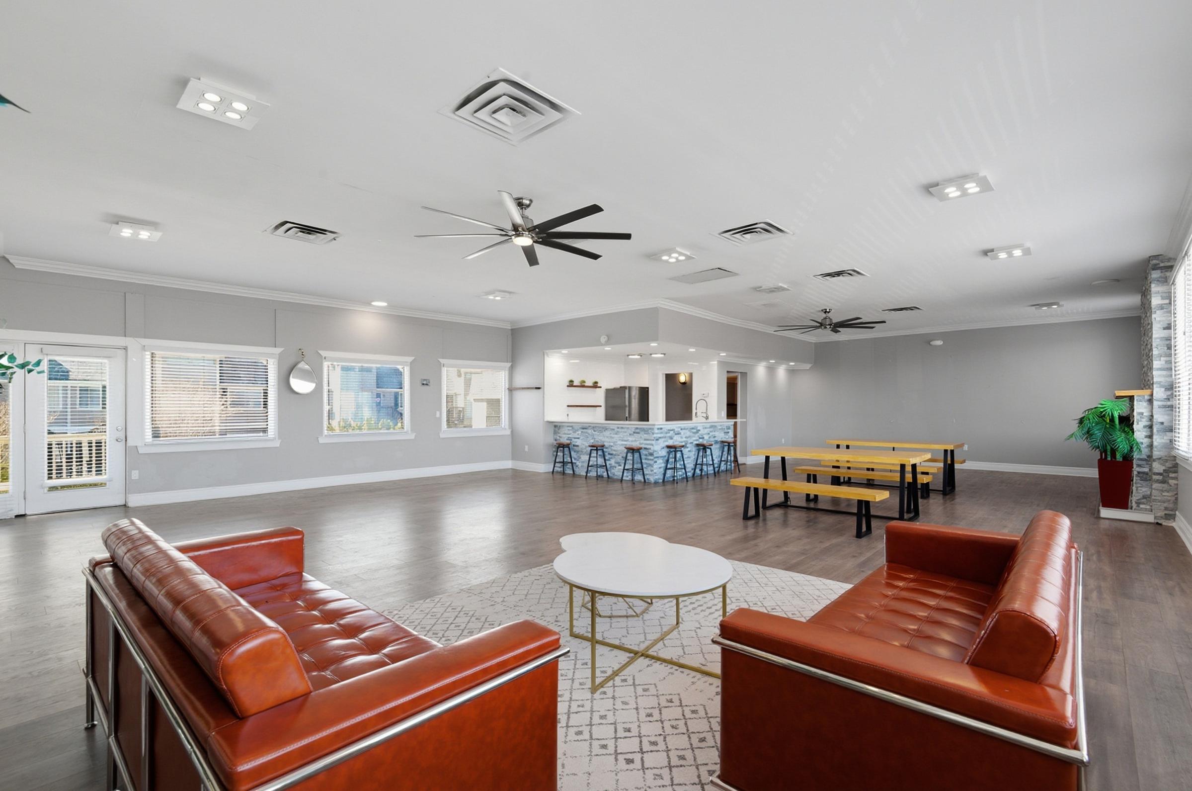 A spacious community lounge featuring modern furniture, including brown leather sofas, a round coffee table, and a large dining area with long tables. The room is brightly lit with ceiling fans and large windows, creating an inviting atmosphere for gatherings or relaxation.