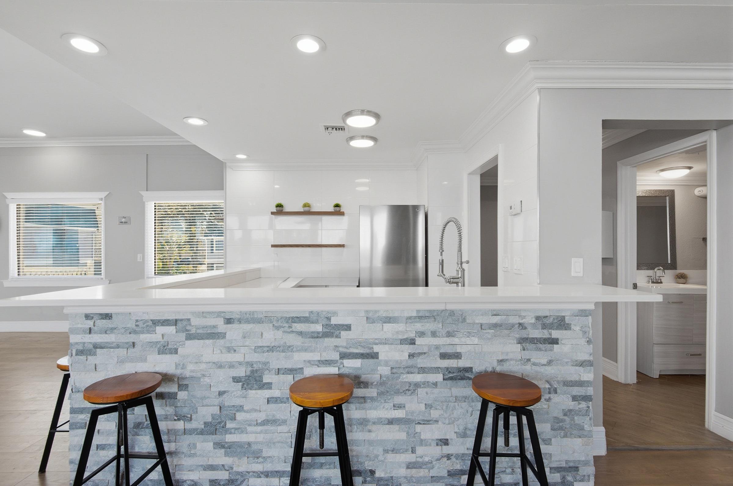 Modern kitchen featuring a breakfast bar with three wooden stools, a sleek countertop, stainless steel appliances, and bright overhead lights. The design showcases a mix of white cabinetry and gray stone accents, creating a clean and inviting atmosphere. Large windows provide natural light.