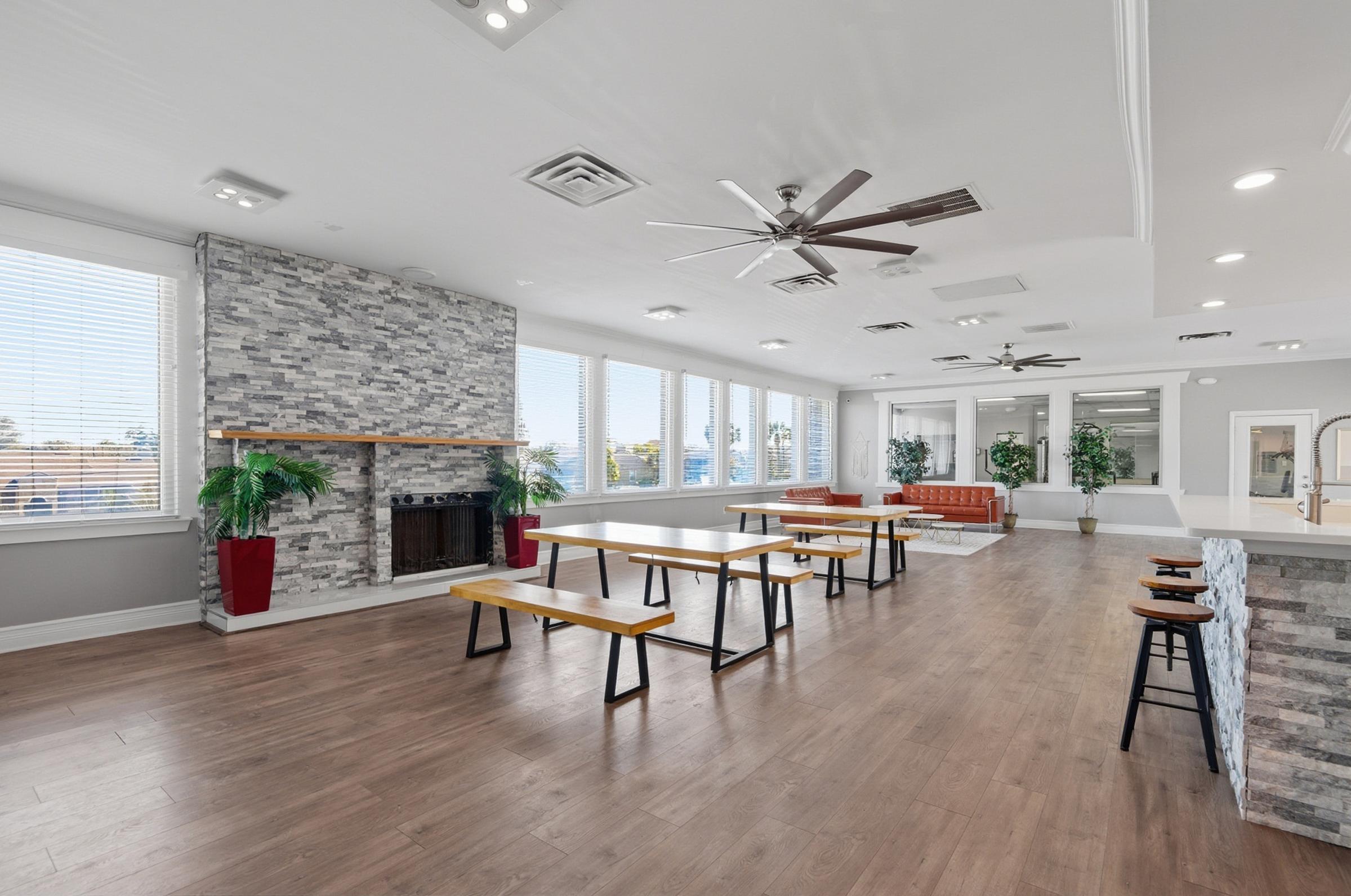 A spacious interior featuring large windows providing natural light. There's a stone fireplace on one side, wooden benches and tables in the center, and a cozy seating area with a couch. Potted plants add a touch of greenery, creating an inviting atmosphere.