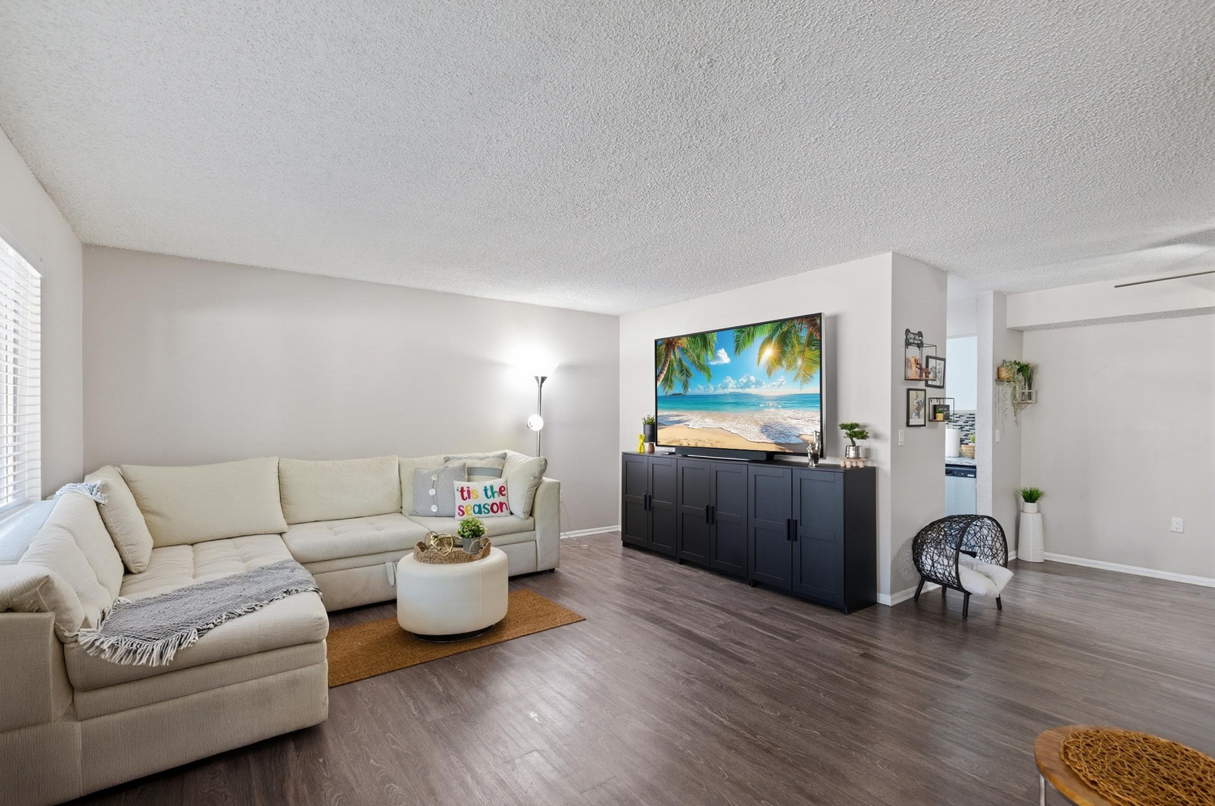 A cozy living room featuring a light-colored sectional sofa, a round coffee table, and a large TV mounted on a black entertainment unit. The space is well-lit with natural light from a window, complemented by decorative elements like plants and a woven basket. The flooring is a warm, wood-like laminate.