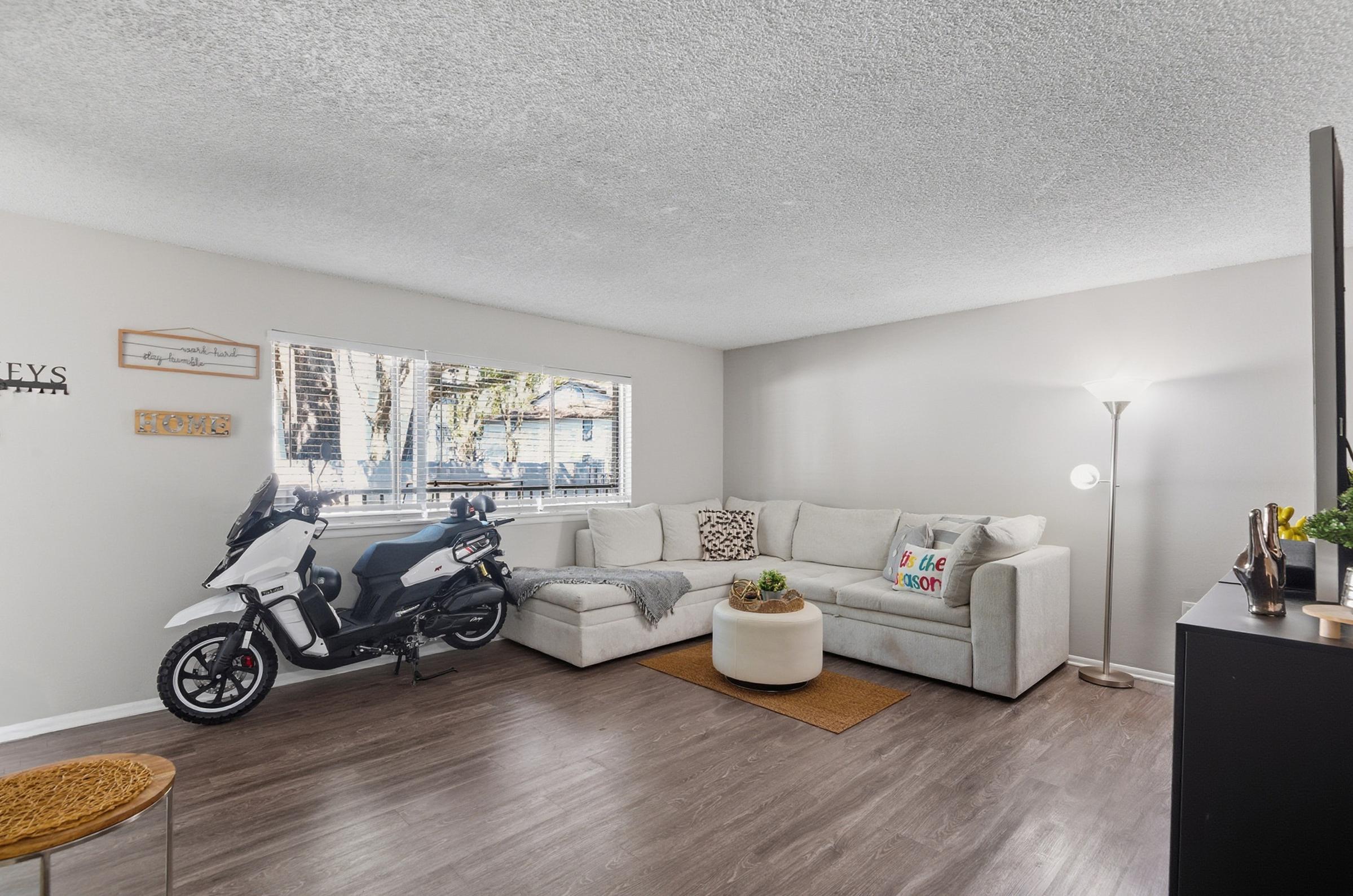 A modern living room featuring a light gray sectional sofa, a round white coffee table on a brown rug, and a floor lamp. A black scooter is parked next to the sofa. There are large windows with natural light and some greenery visible outside. The décor includes decorative pillows and wall art.
