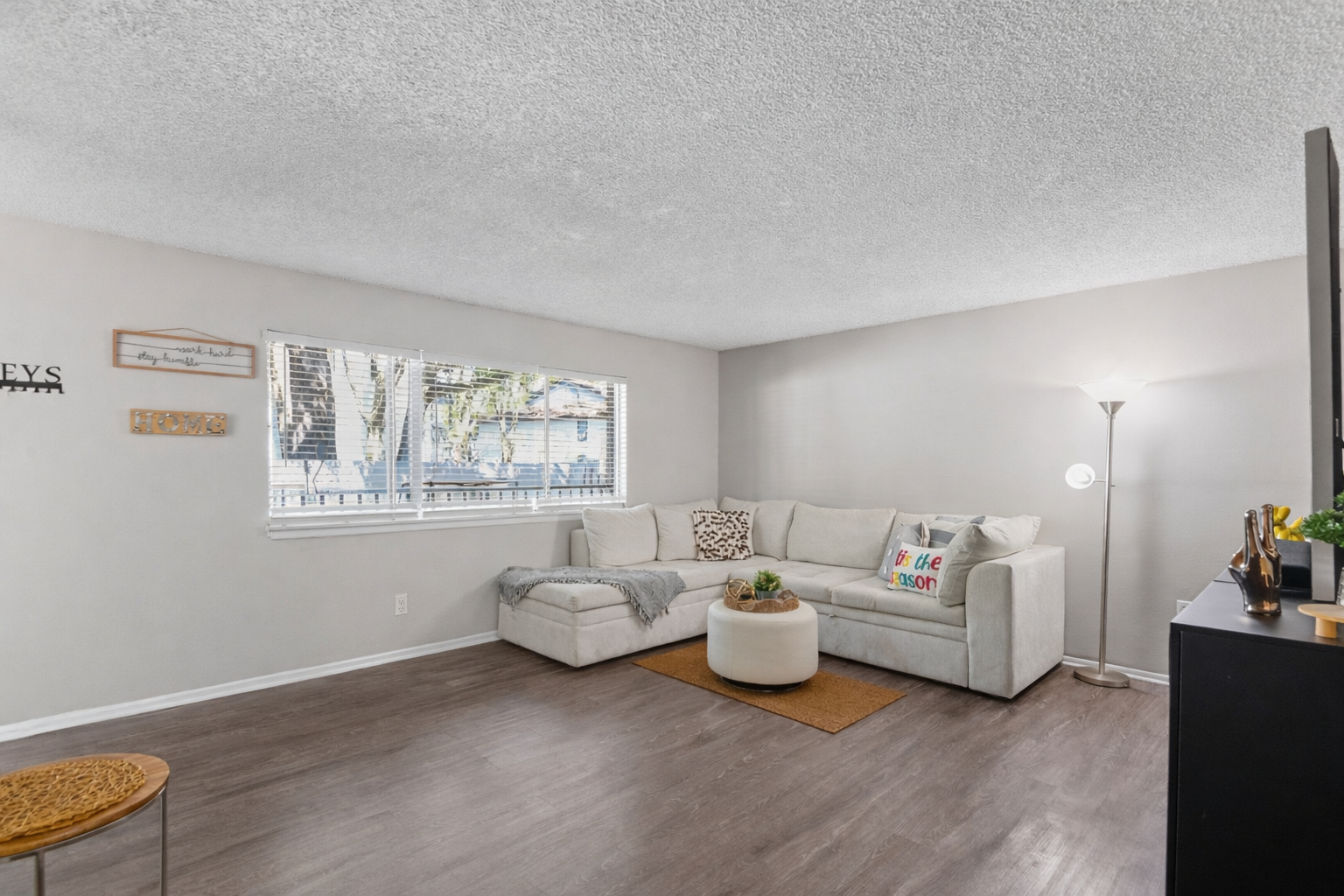 A cozy living room featuring a light-colored sectional sofa adorned with decorative pillows, a round coffee table on a beige rug, and a floor lamp. Natural light flows through a window with white blinds. Simple wall decor adds a homey touch to the space.