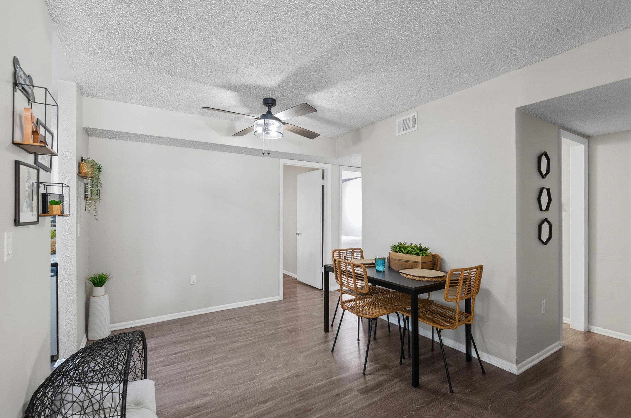 A cozy, modern dining area featuring a small table with four woven chairs. The light-colored walls complement the sleek dark wood flooring. A ceiling fan provides ventilation. Decorative wall accents include a plant and geometric shapes, adding a touch of warmth to the space.