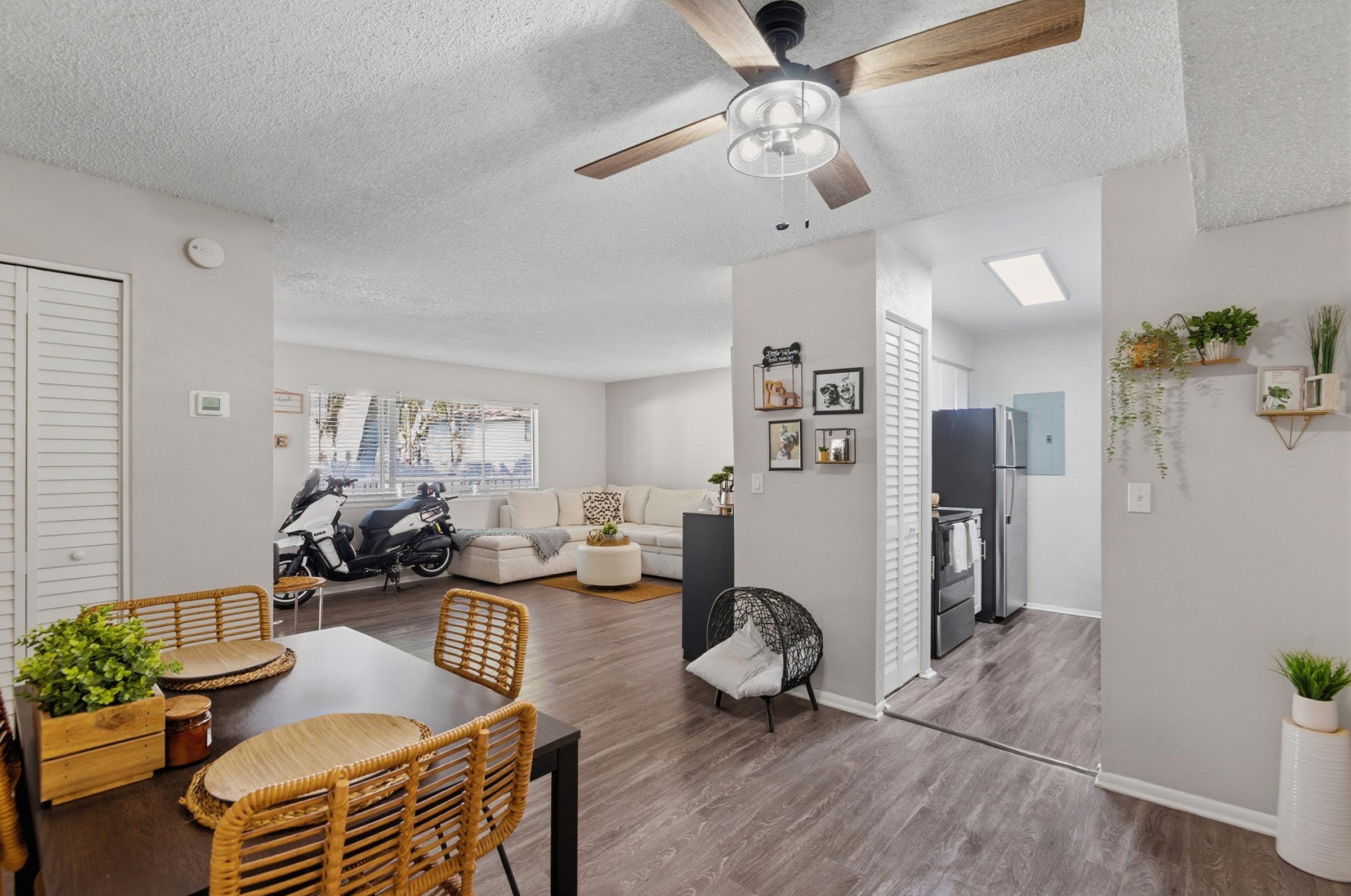 Interior view of a modern apartment featuring an open layout. The dining area has a wooden table with woven chairs. The living space includes a gray sofa and a motorcycle parked near the window. A modern kitchen is visible in the background, with a refrigerator and decorative plants on the walls.
