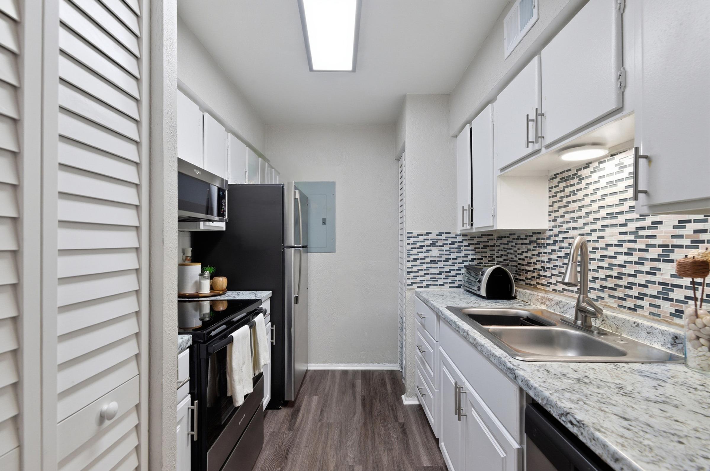 Modern kitchen featuring white cabinetry, a stainless steel refrigerator, and a black stove. The countertops are made of granite, and a backsplash with blue mosaic tiles adds a stylish touch. The space is well-lit with overhead lighting and has a clean, contemporary design.