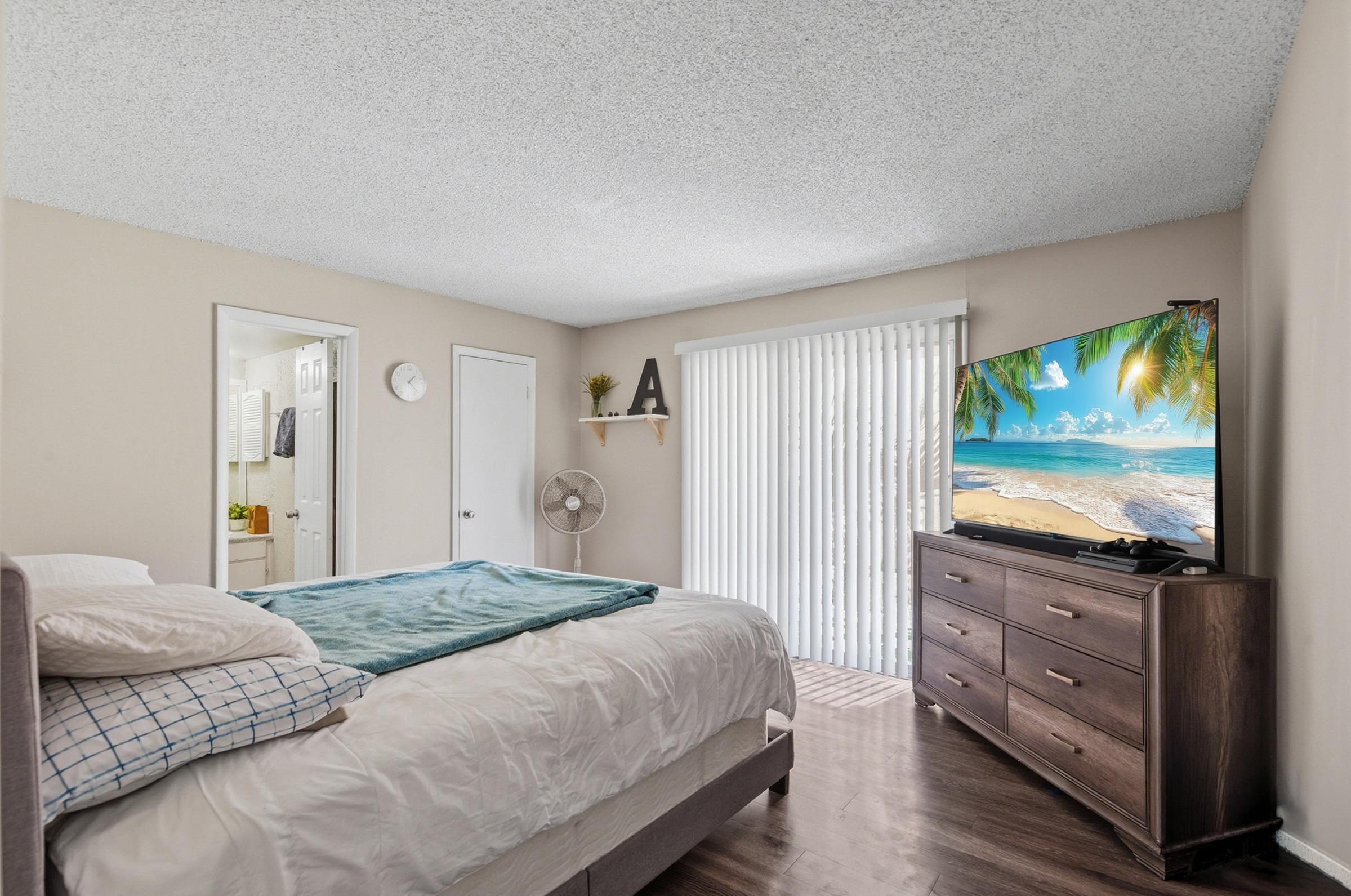 A cozy bedroom featuring a queen-sized bed with a light blue blanket, a wooden dresser with a TV displaying a beach scene, a ceiling fan, and a sliding door that opens to a sunny outdoor view. The walls are painted light beige, creating a warm and inviting atmosphere.
