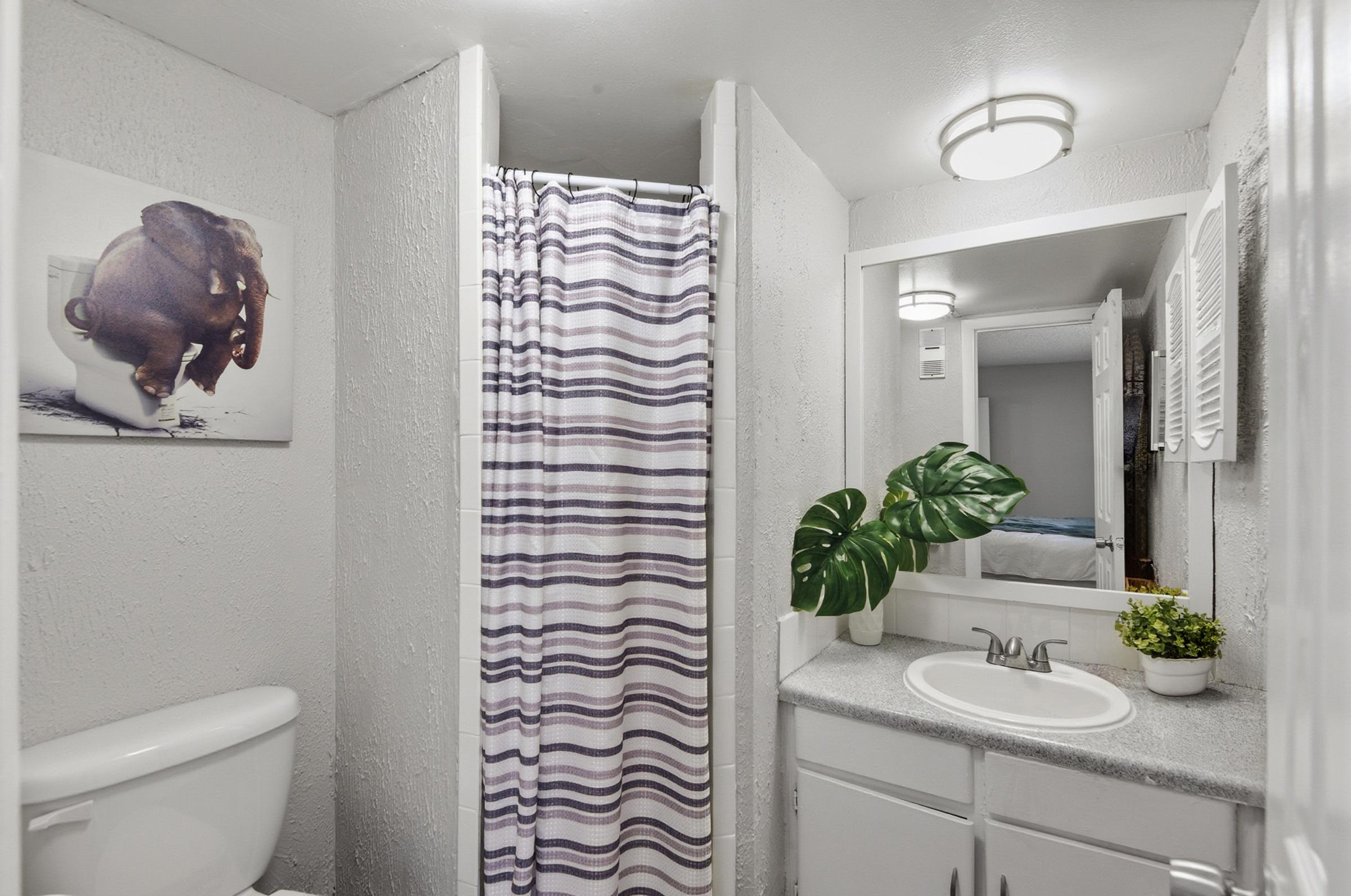 A small bathroom featuring a toilet, a shower with a striped curtain, and a sink with a plant. The wall has a humorous art piece of an elephant sitting on a toilet. Soft lighting from a ceiling fixture and a mirror enhance the bright, clean space. The decor includes a tropical plant for a fresh touch.