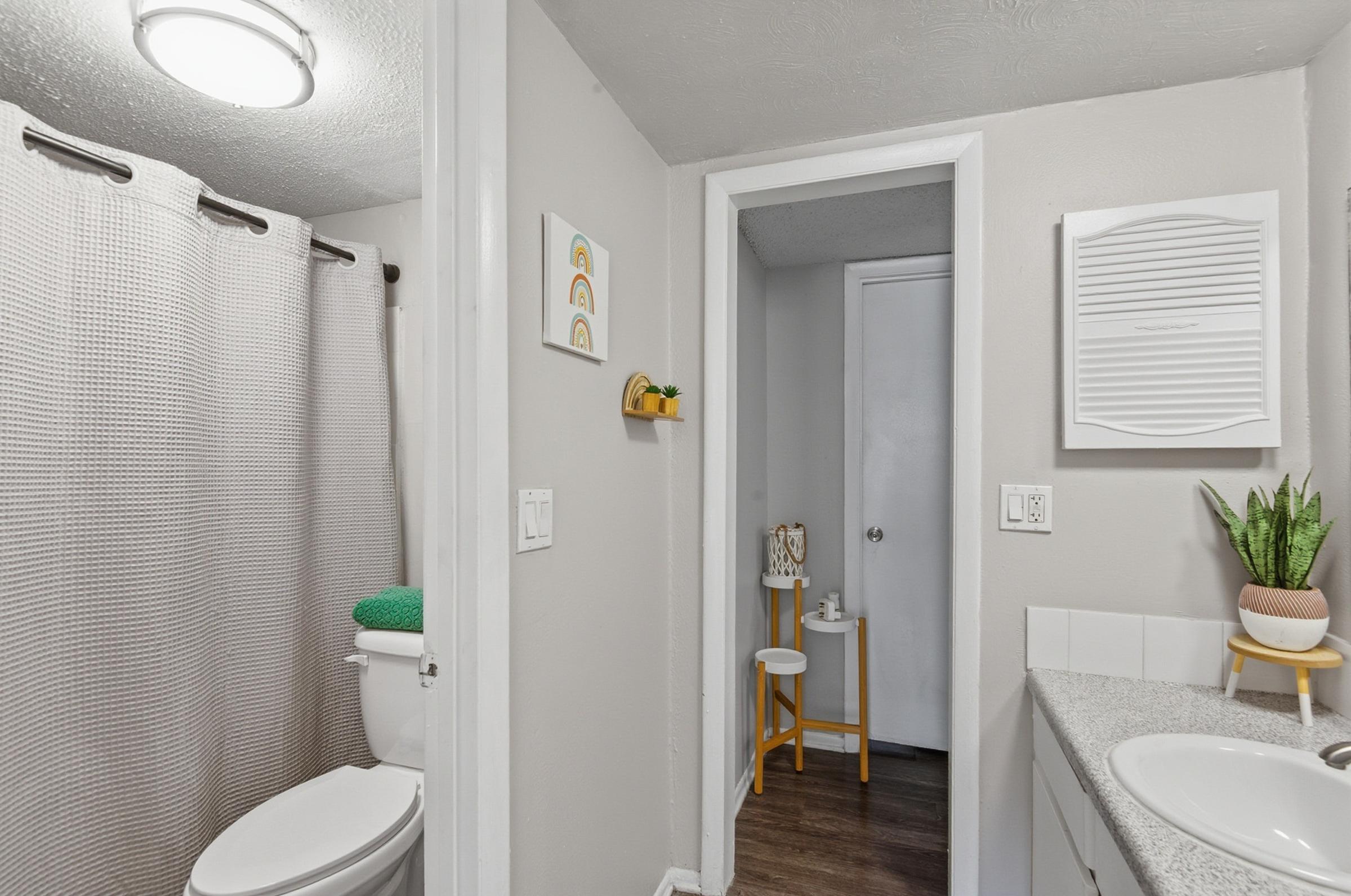 A clean bathroom featuring a shower area with a textured curtain, a toilet, and a sink with light-colored cabinetry. The walls are painted in a soft tone, and there are decorative elements such as wall art and a small plant. A door leads to a separate small room, visible in the background.