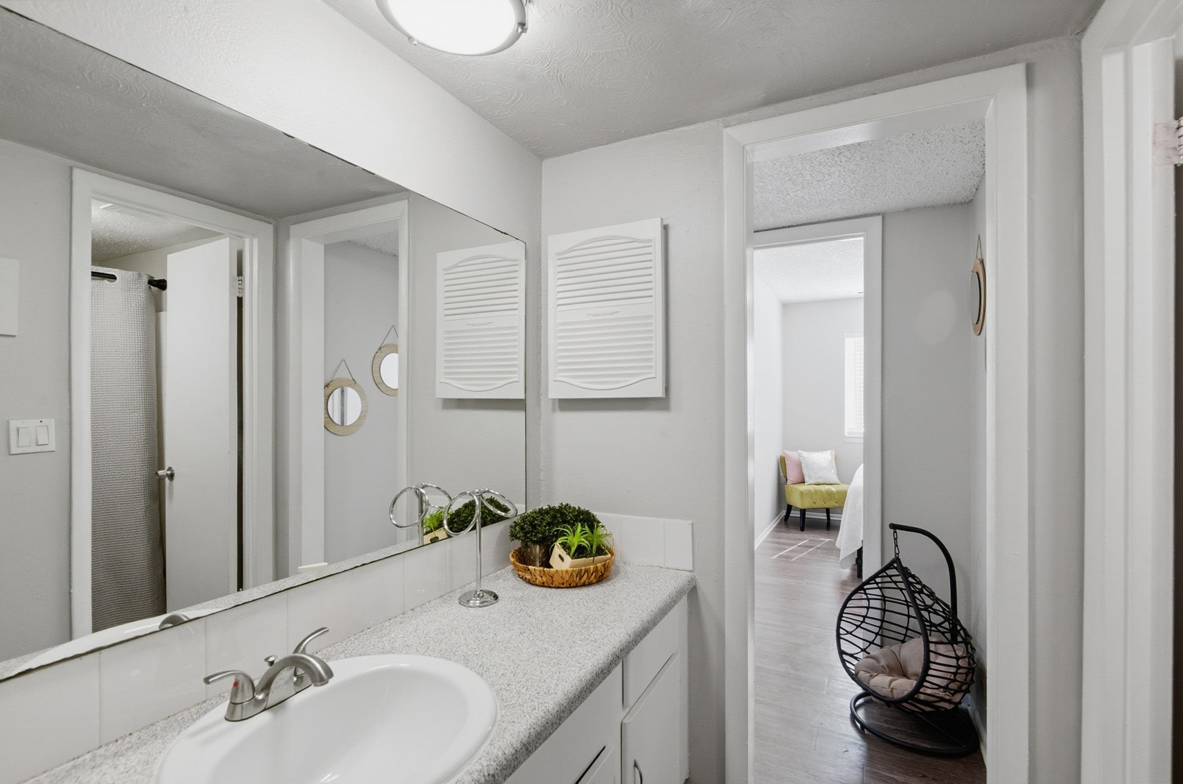 A modern bathroom with a light gray color scheme. It features a white sink with a chrome faucet, a large mirror, and an illuminated ceiling fixture. On the countertop, there is a decorative basket with greenery. A doorway leads to another room, and a cozy seating area is visible in the background.