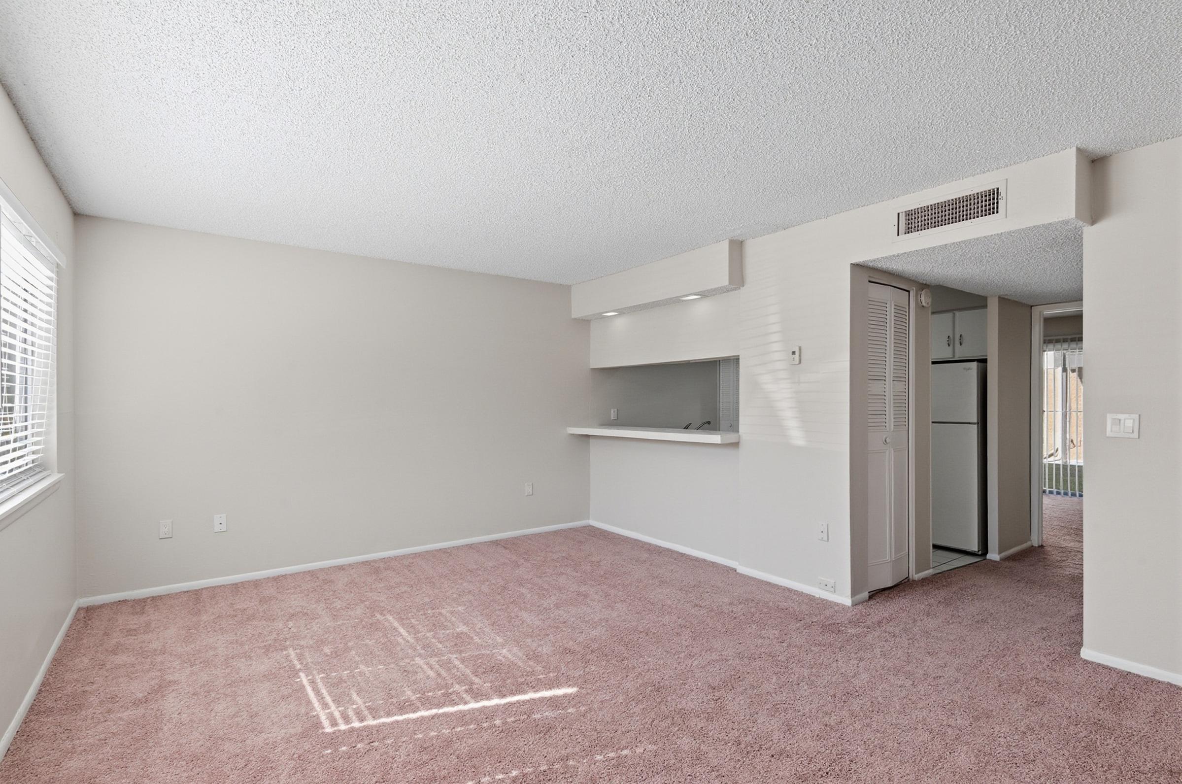A spacious and well-lit living area featuring light-colored walls, plush pink carpet, and a large window with blinds. To the right, an open kitchen area can be seen, along with a refrigerator and a doorway leading to another room. The overall ambiance is warm and inviting.
