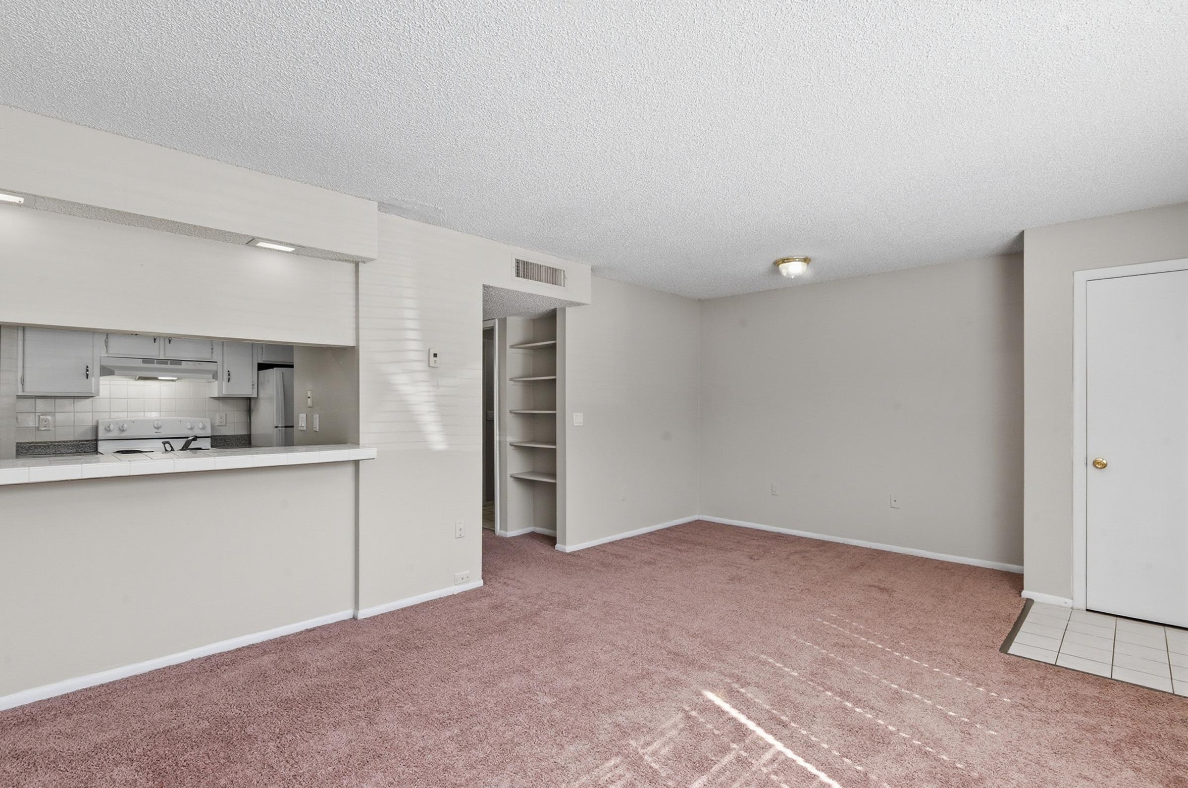 A spacious, well-lit living room with beige walls and light brown carpet. There is an open kitchen area with a counter visible in the background, and a small shelving unit on the right. A door on the right leads to another area, and indirect sunlight filters in through a window, creating a warm ambiance.