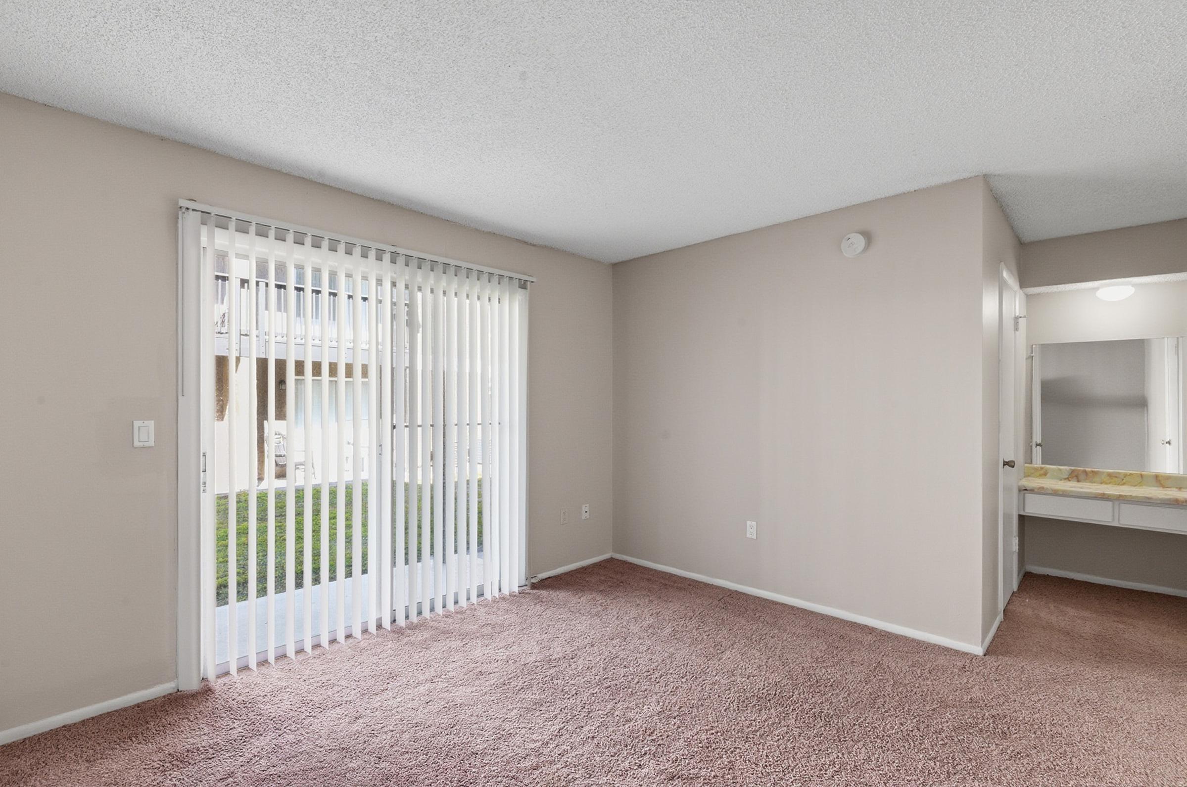 A light and spacious room with beige walls and carpet flooring. It features a sliding glass door with vertical blinds that opens to a grassy outdoor area. A small vanity area is visible on the right side, with a mirror and counter space, contributing to a bright and inviting atmosphere.
