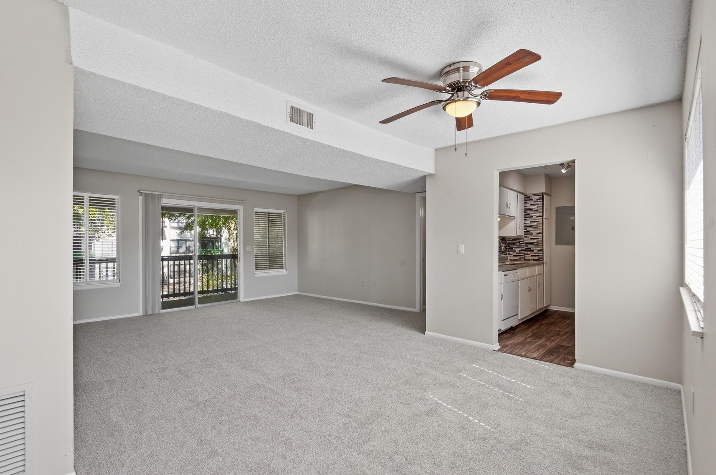 A spacious living room features light-colored walls and plush carpeting, with a ceiling fan and large windows offering natural light. The room opens to a small kitchen area in the background, showcasing modern cabinetry and a stylish backsplash. The overall atmosphere is bright and inviting.
