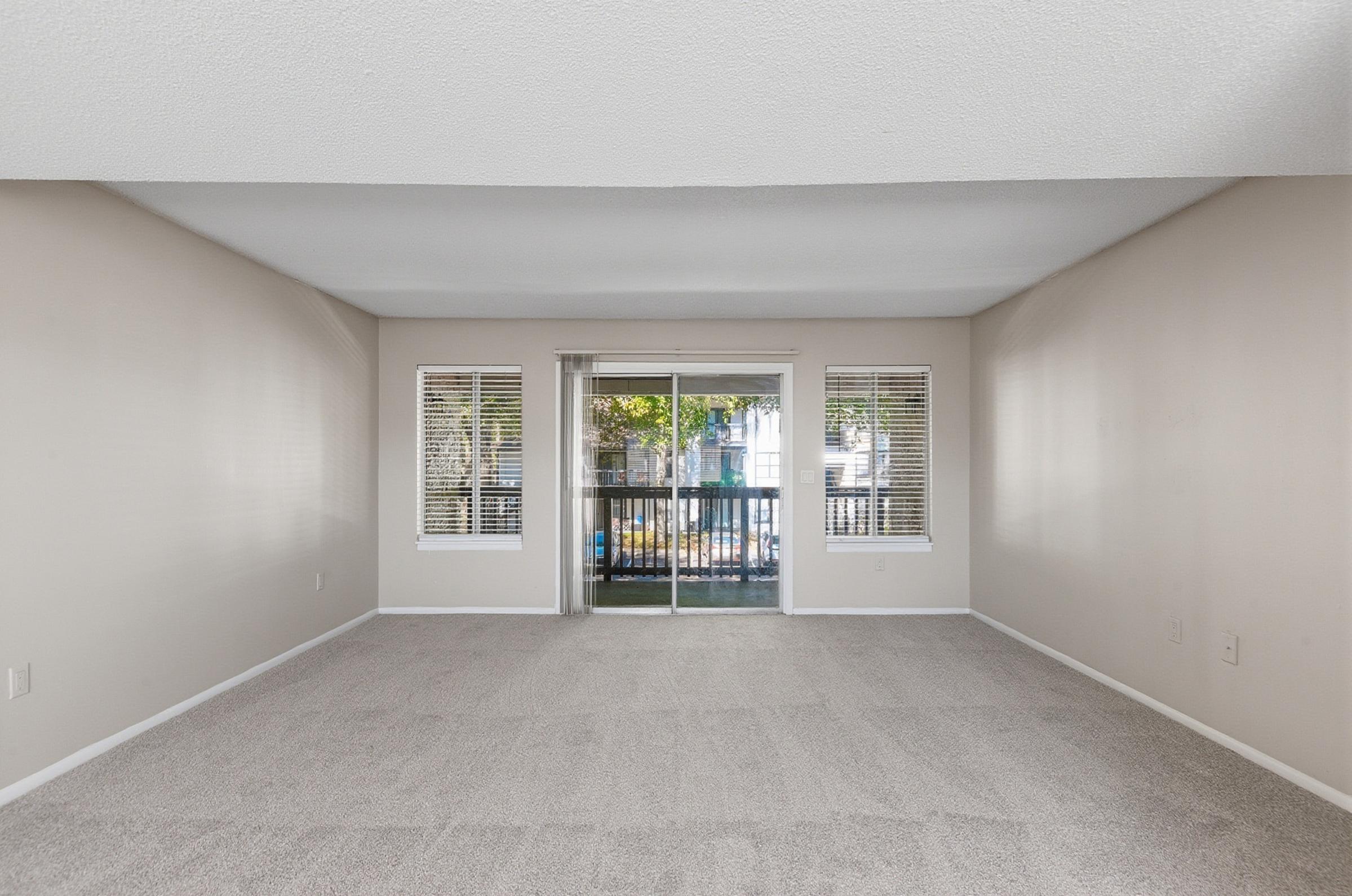 A spacious, empty living room with light-colored walls and plush carpet. Large windows with white blinds provide natural light and a view of a balcony. The room has a clean, minimalist aesthetic, ideal for personal decoration.