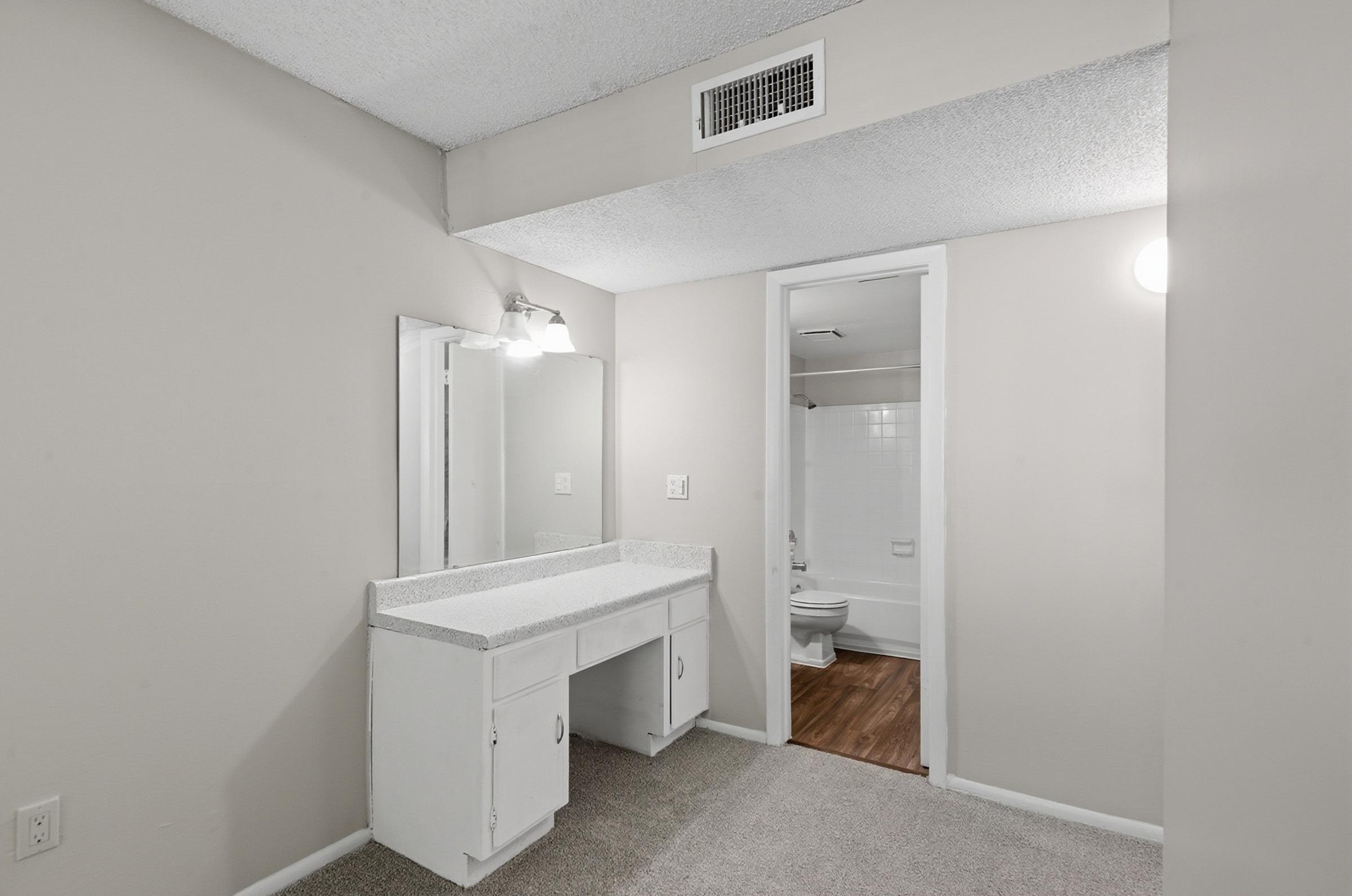 A clean and simple bathroom vanity area with a mirror, white countertop, and light fixture. The walls are painted a light color, and there is a doorway leading to a bathroom with a toilet visible in the background. The floor features a wood-like finish, and the carpet is a neutral color.