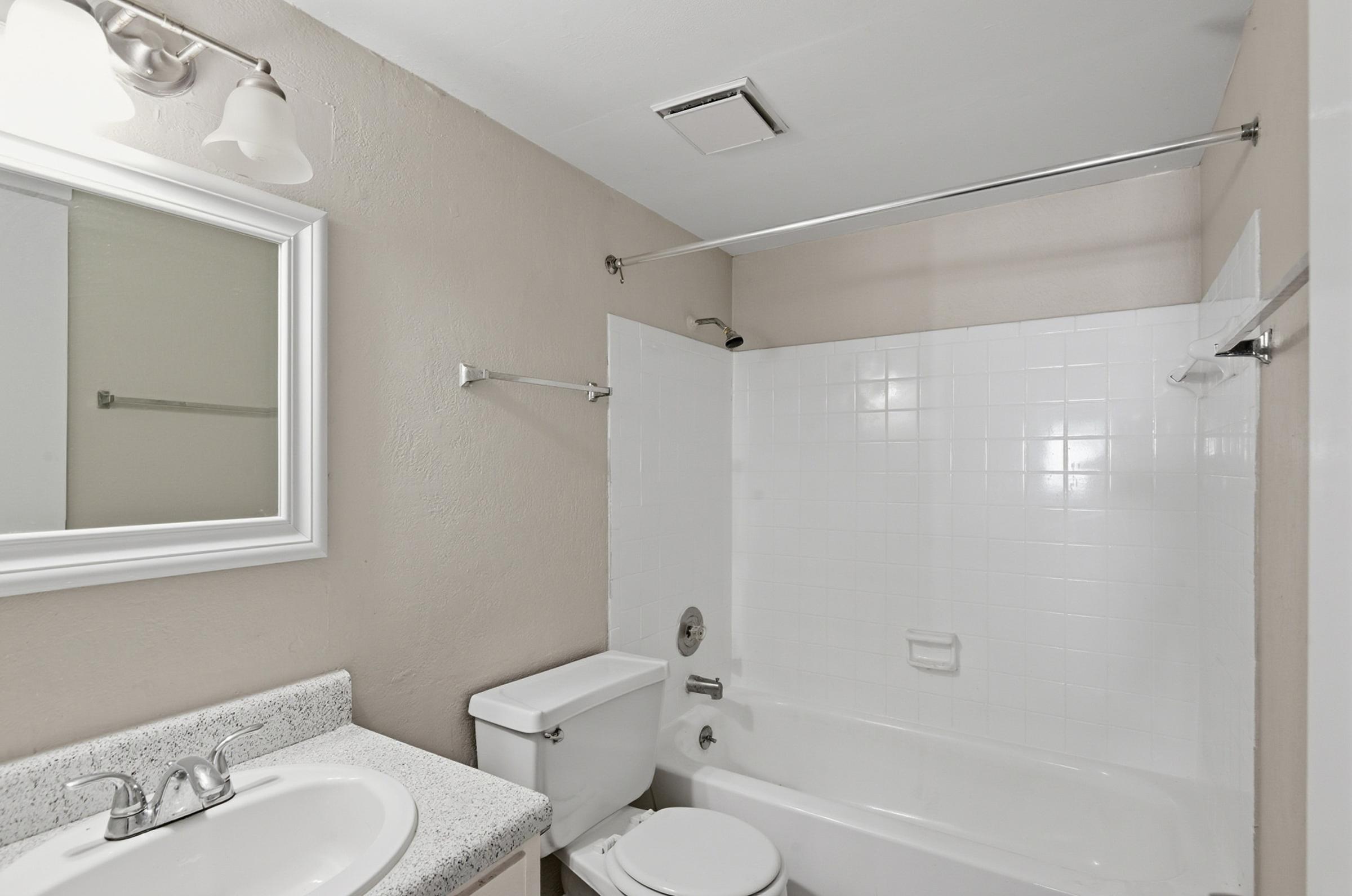 A clean and modern bathroom featuring a white bathtub with a shower curtain, a toilet, a sink with a granite countertop, and a wall-mounted mirror. The walls are painted in a soft beige, and there is a ceiling light fixture providing adequate illumination.