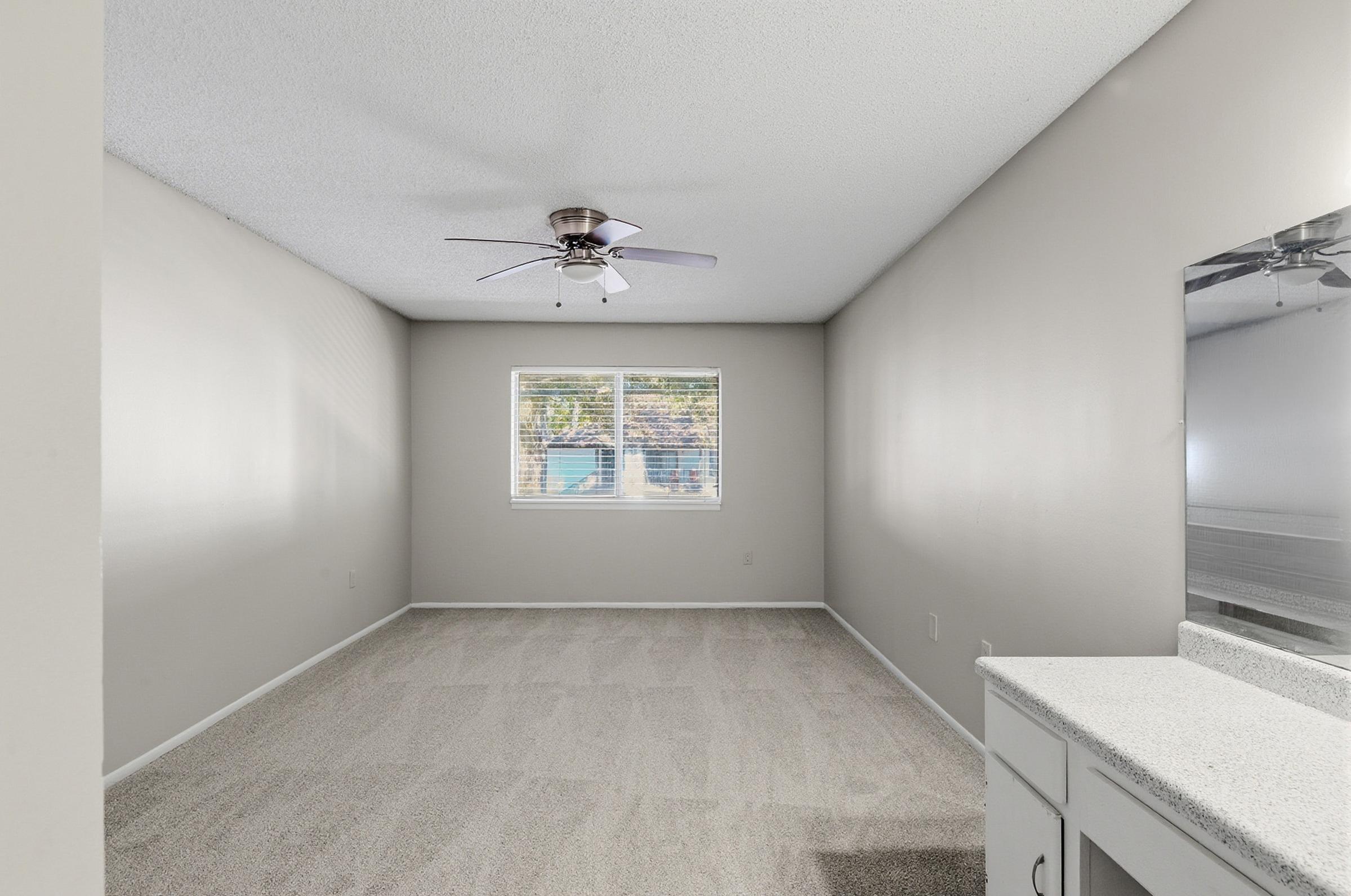 A spacious, empty room with light gray walls and a ceiling fan. There is a window that lets in natural light, and the floor is covered in light-colored carpeting. A small vanity with a countertop is visible on one side of the room, creating a bright and open atmosphere.