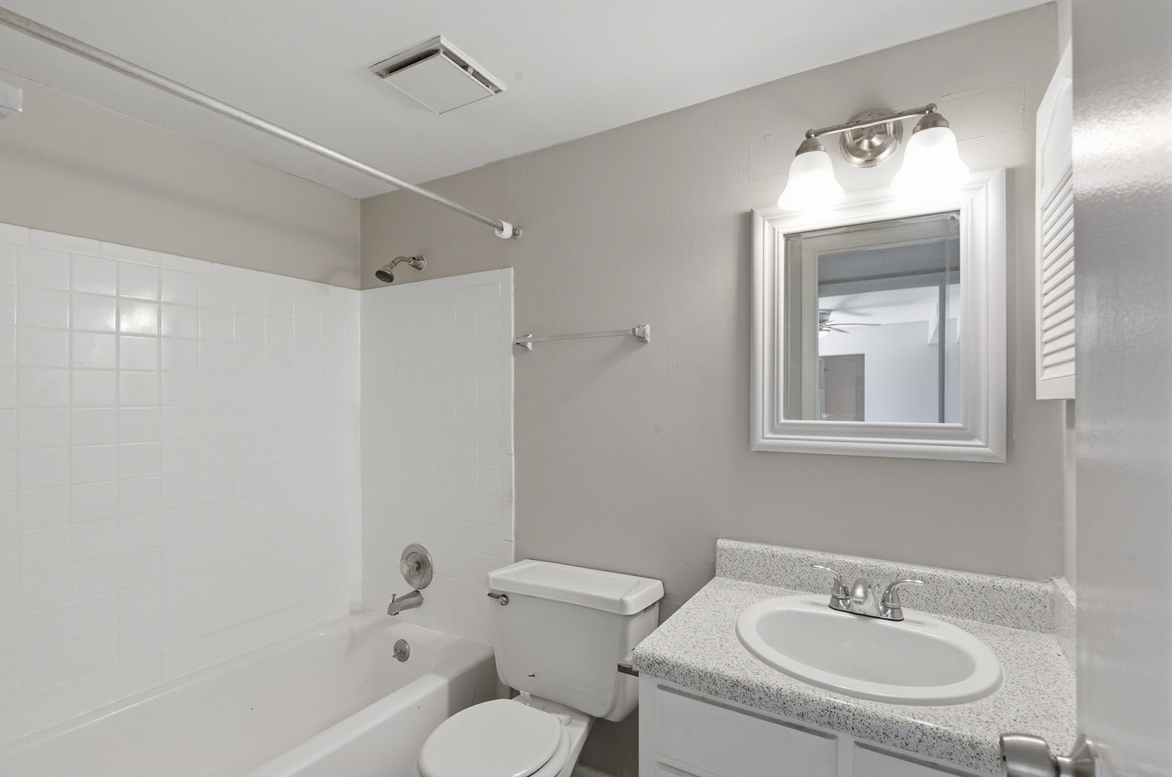 A clean and modern bathroom featuring a white tiled bathtub and shower, a toilet, and a granite countertop vanity with a sink. A mirror is mounted above the vanity, and there is a light fixture above the mirror. The walls are painted in a neutral color, creating a spacious feel.