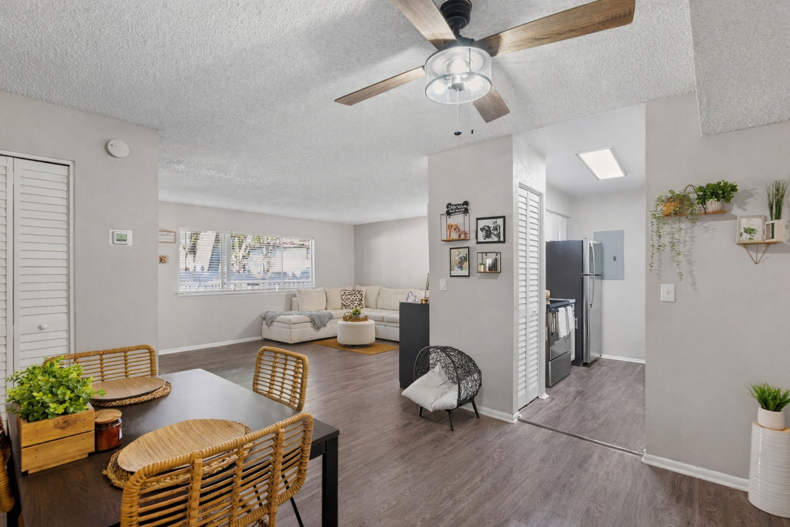 A bright, modern living space featuring an open floor plan. The room includes a dining area with a table and woven chairs, a cozy seating area with a light-colored sofa, and a kitchen nook with a refrigerator. Natural light flows in through windows, highlighting the neutral tones and decorative accents.