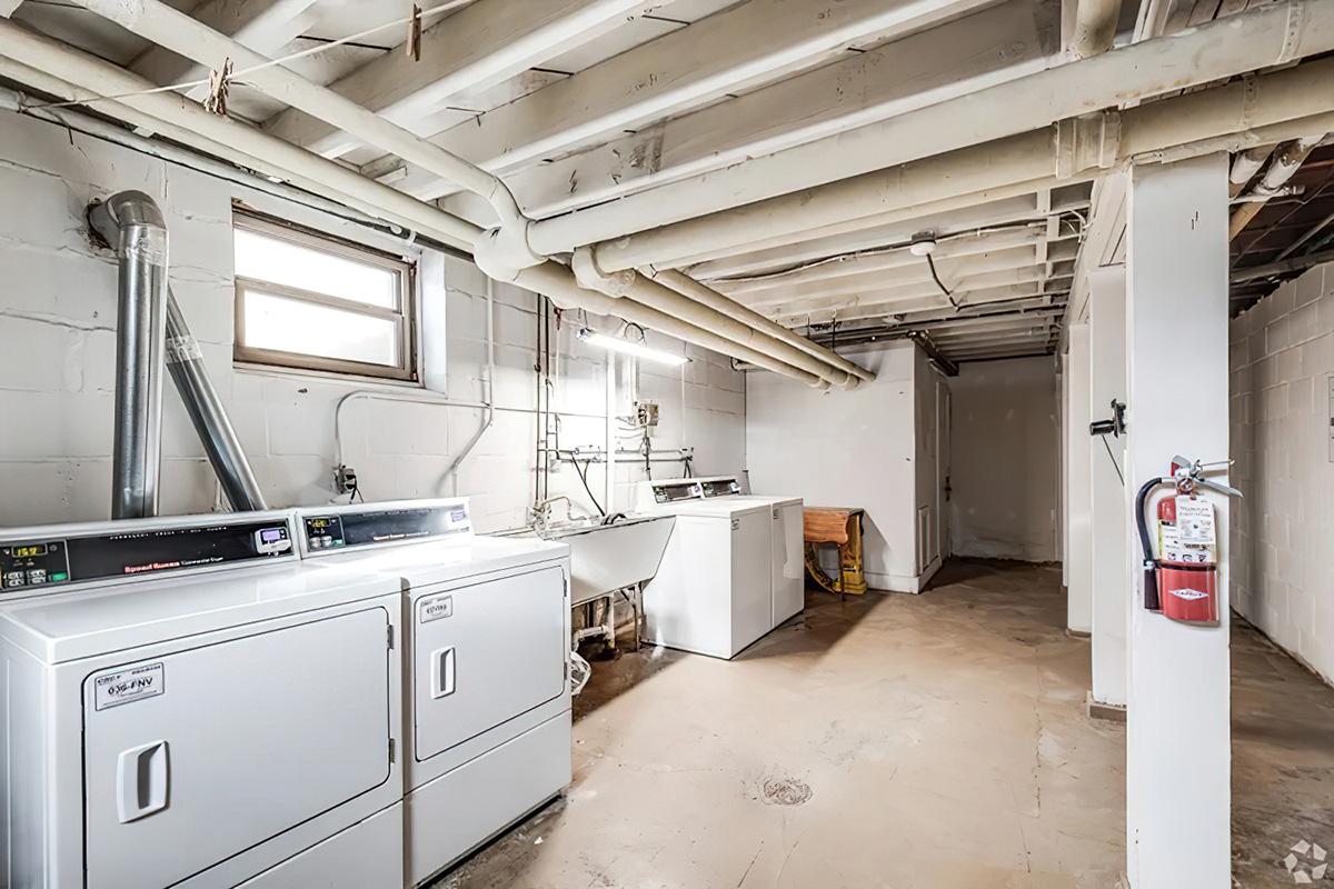 A clean, unfinished basement laundry room featuring two white washing machines, a utility sink, and a dryer. The space has exposed ceiling beams and pipes, with concrete floors and white walls. A small window provides natural light, and a fire extinguisher is mounted on the wall.