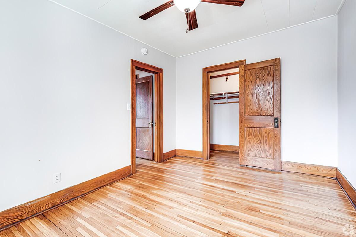 A spacious, light-colored room with wooden flooring, featuring two wooden doors leading to adjacent spaces. The walls are painted white, and there is a ceiling fan in the center. One door opens to a closet with hanging space, and the overall ambiance is clean and uncluttered.