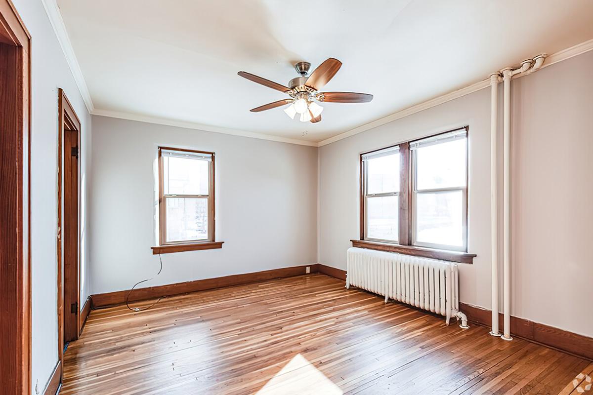 A well-lit, empty room with wooden floors, featuring a ceiling fan and two windows on one wall. There is a radiator along the opposite wall, and light beige walls enhance the spacious appearance. Natural light streams in, creating a warm ambiance.