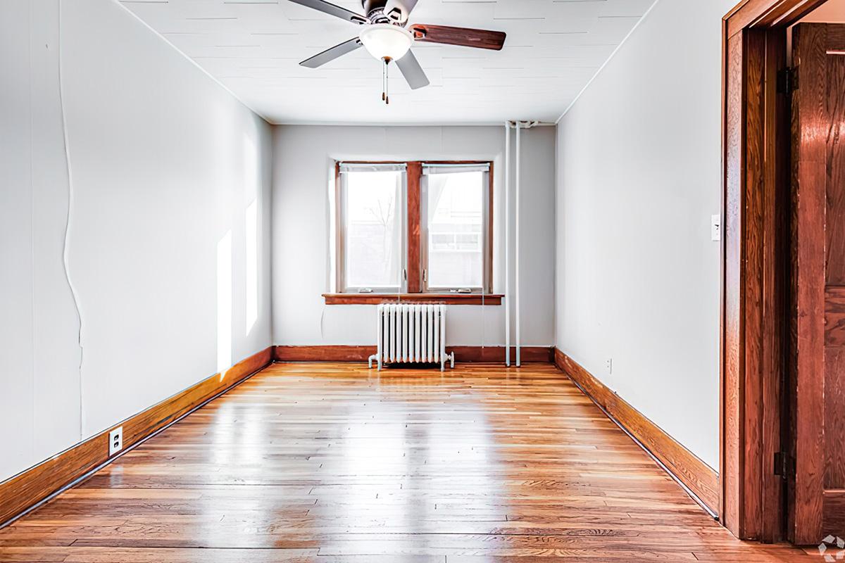 A spacious, empty room featuring light hardwood floors, a ceiling fan, and two windows providing natural light. There is a radiator beneath the windows and wooden trim around the doors and windows, giving the space a modern yet warm feel. The walls are painted in a light color, enhancing the brightness.
