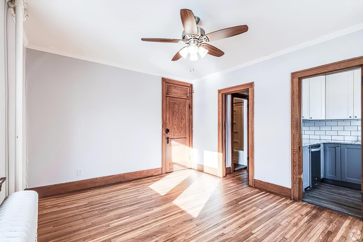A well-lit room featuring hardwood flooring, a ceiling fan with light, and several doorways leading to other areas. The walls are painted in a neutral color, and there are windows allowing natural light to brighten the space. A glimpse of a kitchen area can be seen through one of the doorways.