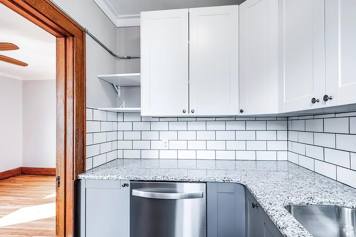 A modern kitchen featuring white cabinets, a granite countertop with a grey and white tiled backsplash, and stainless steel appliances. The space includes an open layout with wooden doorways and flooring, providing a bright and inviting atmosphere.