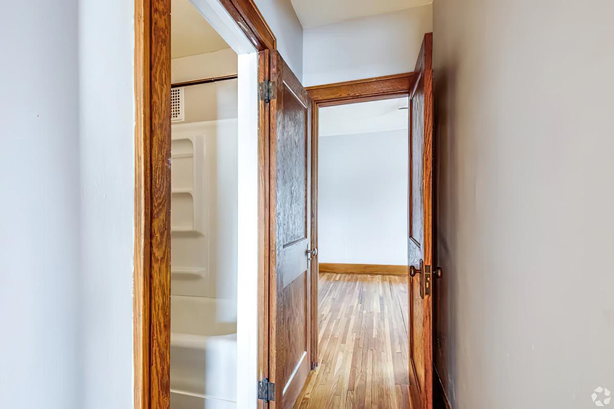 A view of a hallway with doorways leading to different rooms. The left side features an open door leading to a bathroom with a bathtub, while the right side shows an open doorway into another room. The floor is wooden, and the walls are painted in neutral tones. Natural light is visible in the distance.