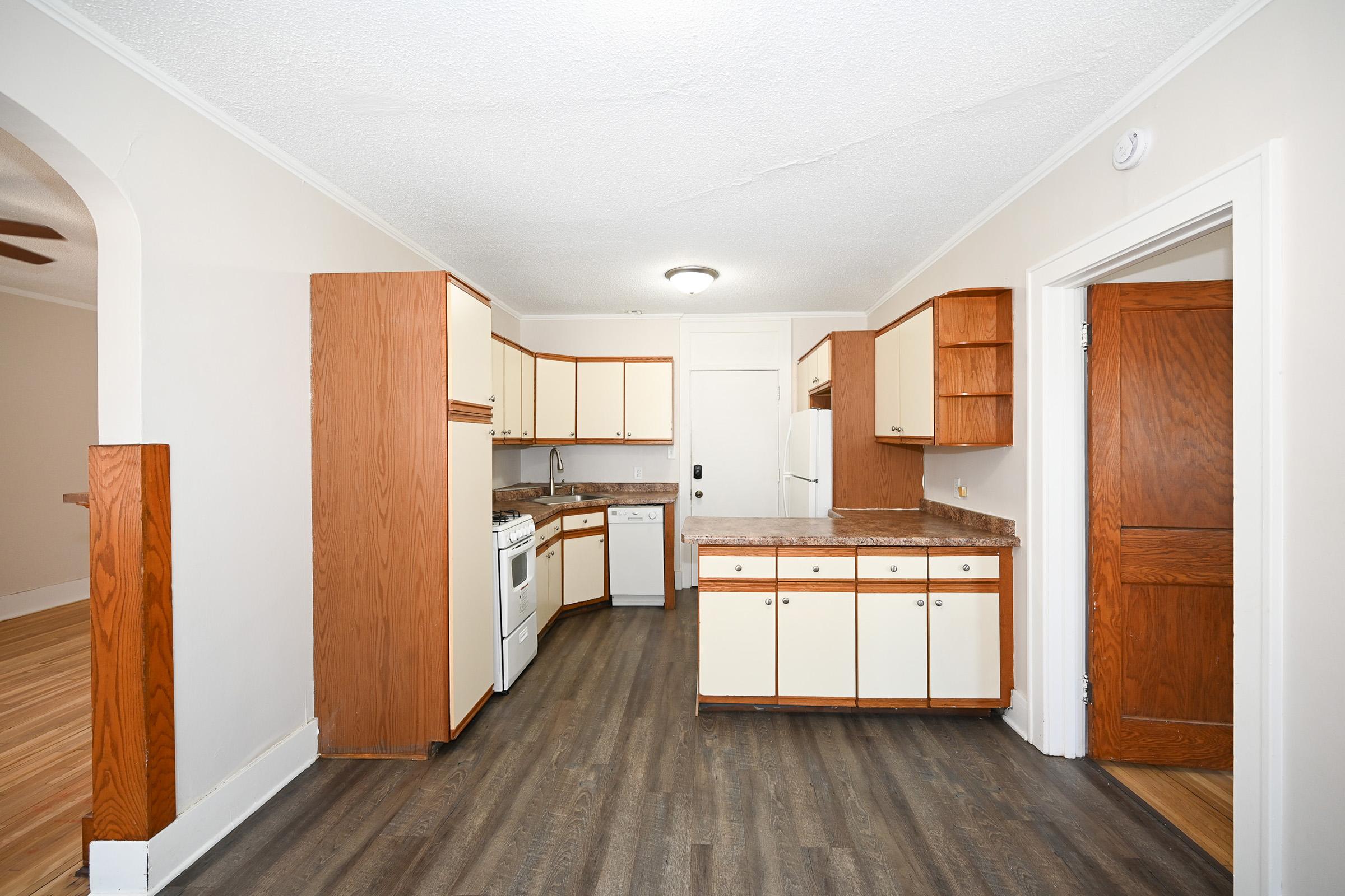 A spacious kitchen with wooden cabinets, white appliances, and a countertop. The layout includes a stove, sink, and dishwasher, with a door leading to another room. The floor is dark laminate, and the walls are painted light beige, giving the space a bright and inviting atmosphere.